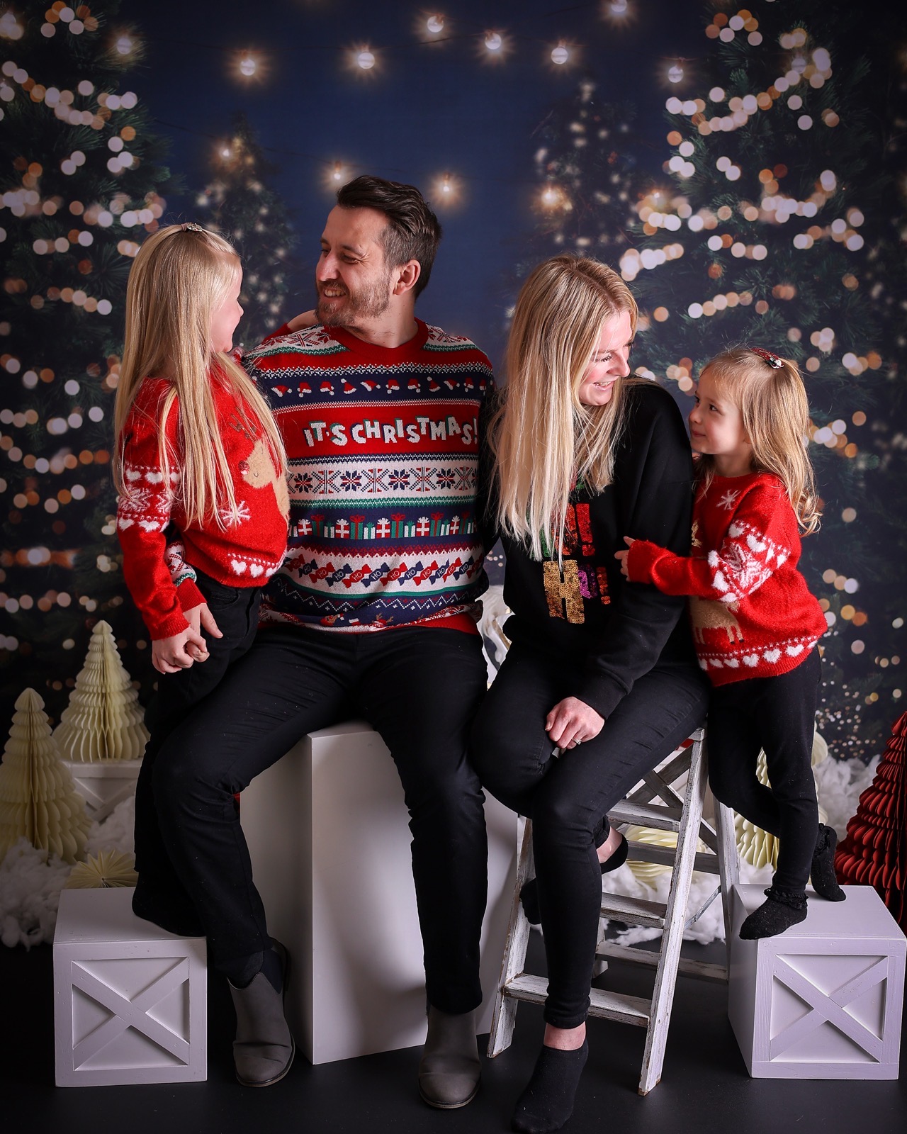 Family of four in matching Christmas jumpers, two young girls with reindeer motifs, dark starry backdrop