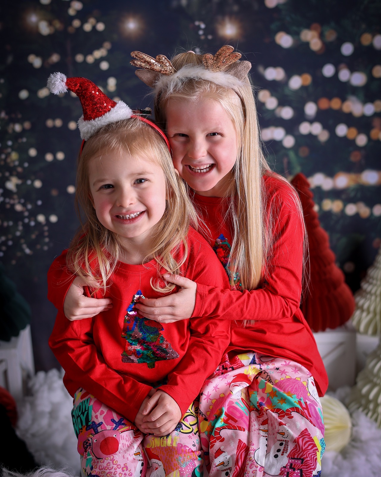 Two young sisters hugging in matching red tops, one in a Santa hat headband and one in reindeer antlers