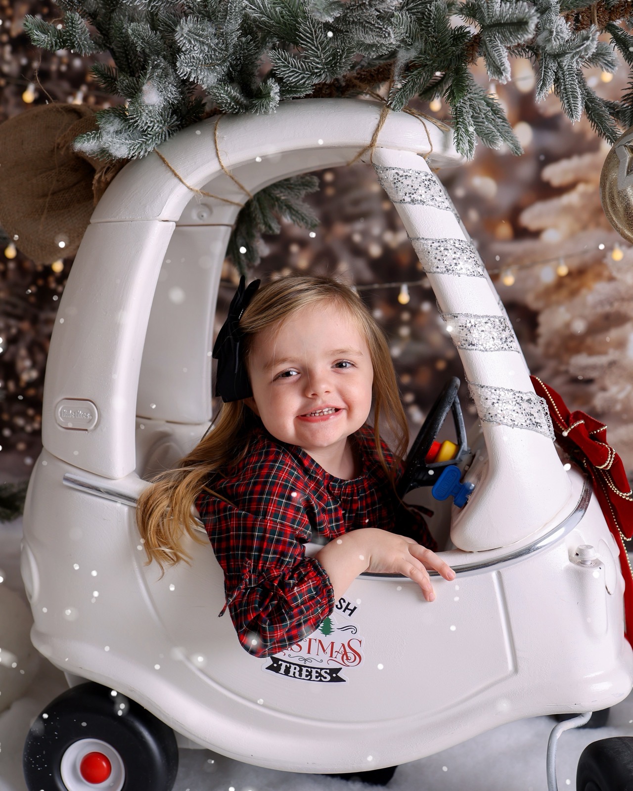 Toddler girl in a red tartan outfit sitting in a toy car under snowy pine branches