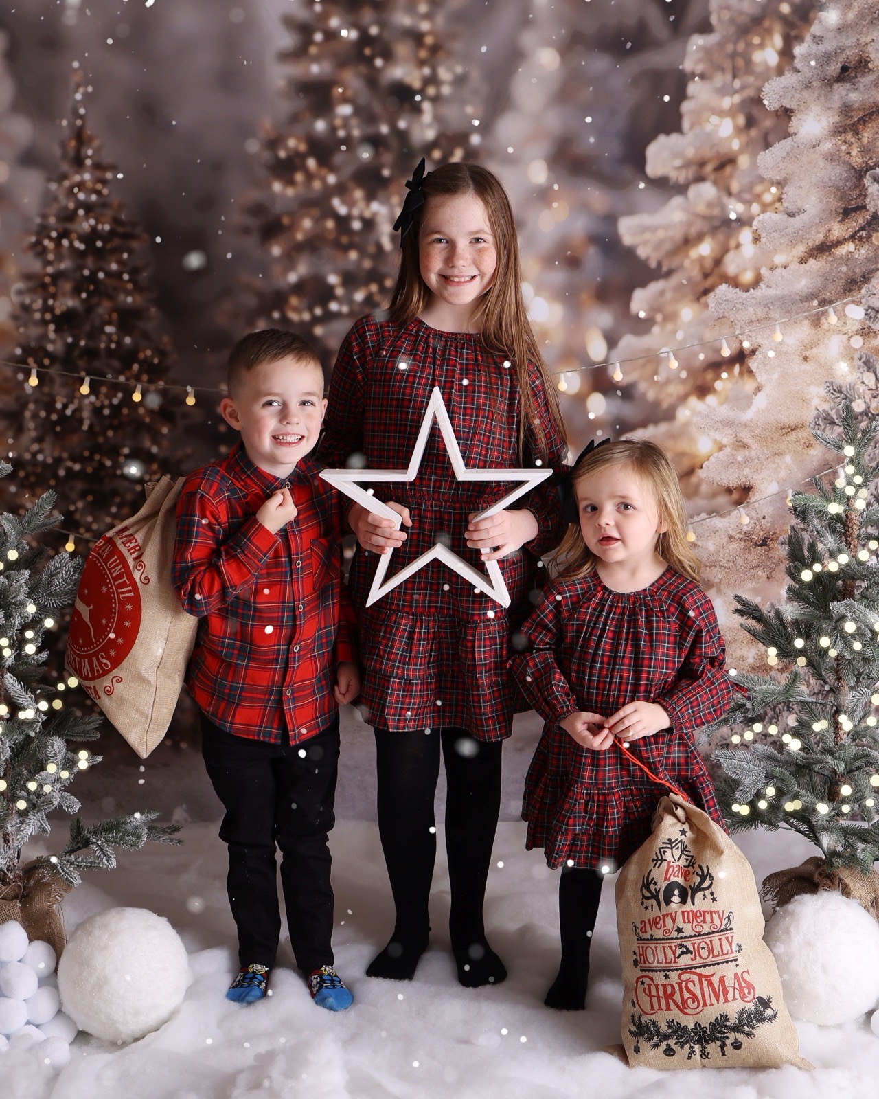 Three siblings in red tartan outfits holding a wooden star and hessian gift sacks, snowy woodland backdrop
