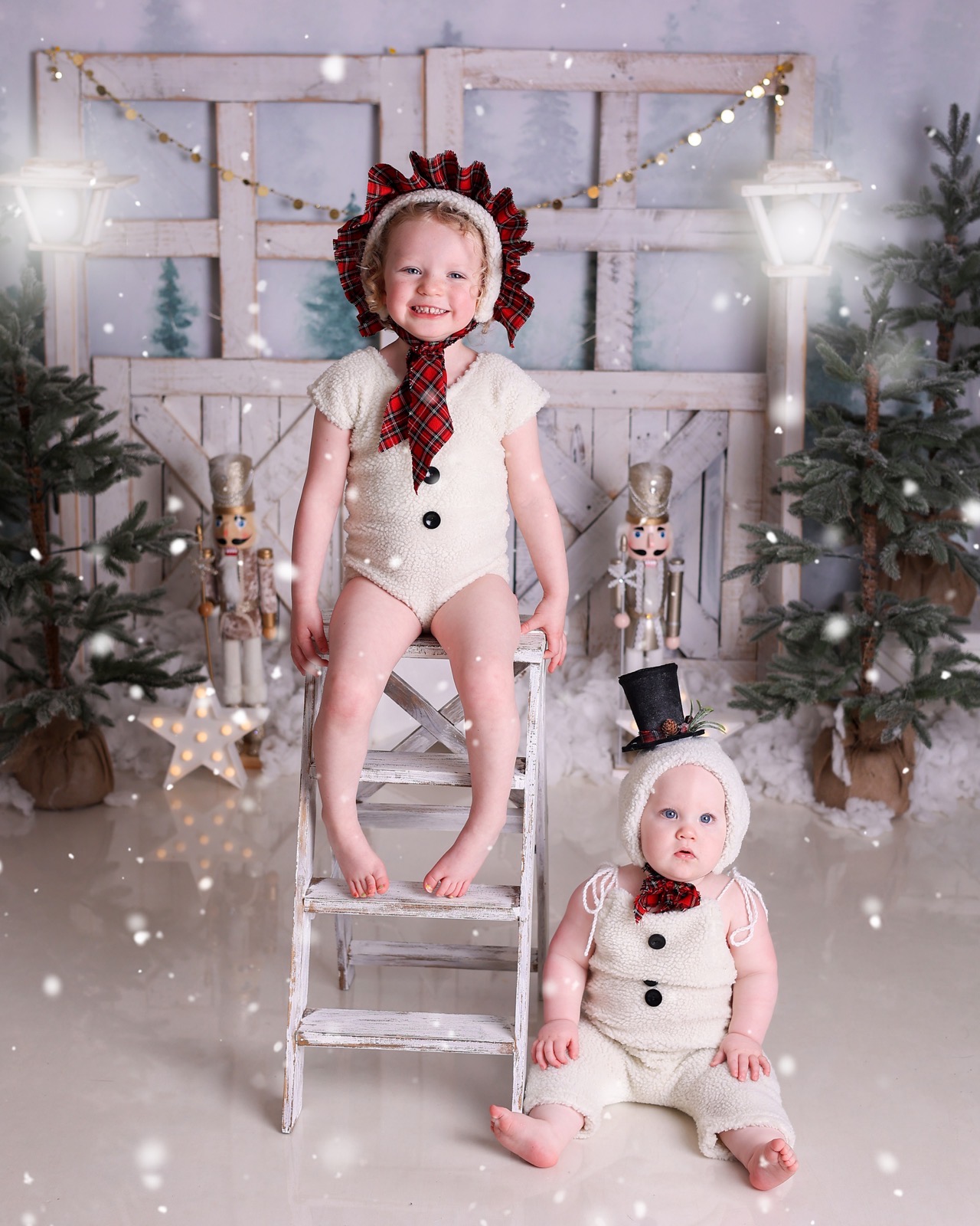 Toddler girl in a snowman bonnet sitting on a small ladder, baby sibling in a matching costume nearby