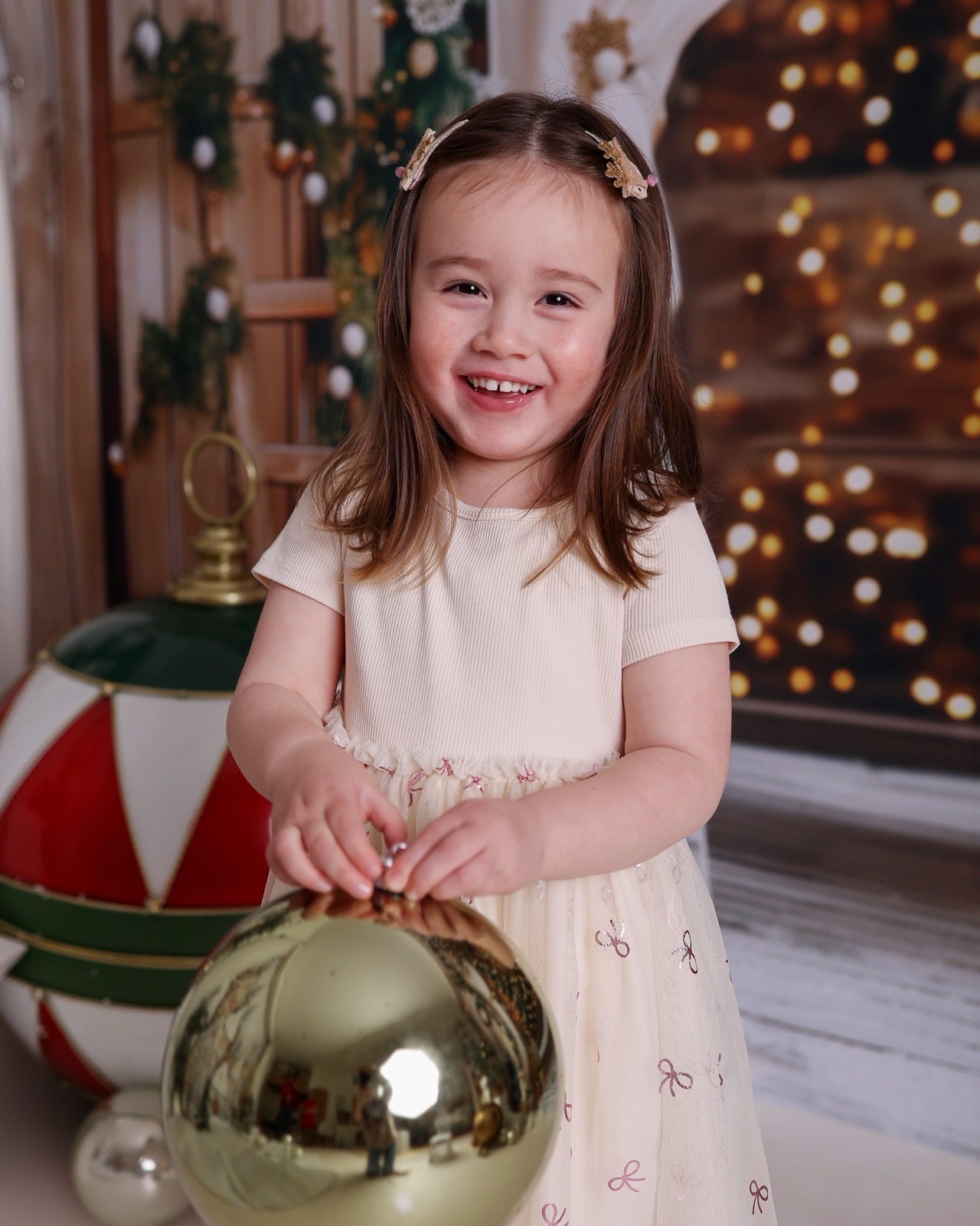 Girl in a cream dress holding a large gold bauble, warm bokeh lights in the background