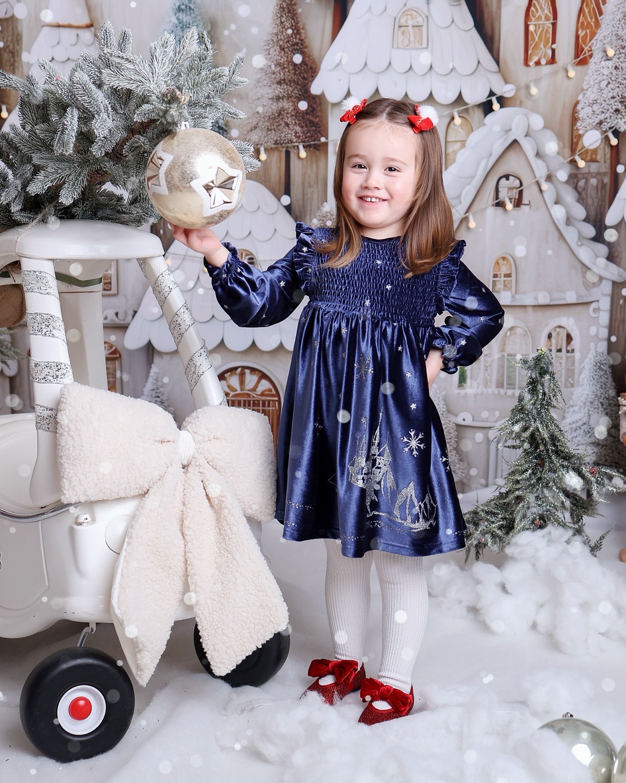Girl in a navy velvet dress with a silver snowflake print, holding a gold ornament beside a toy pram