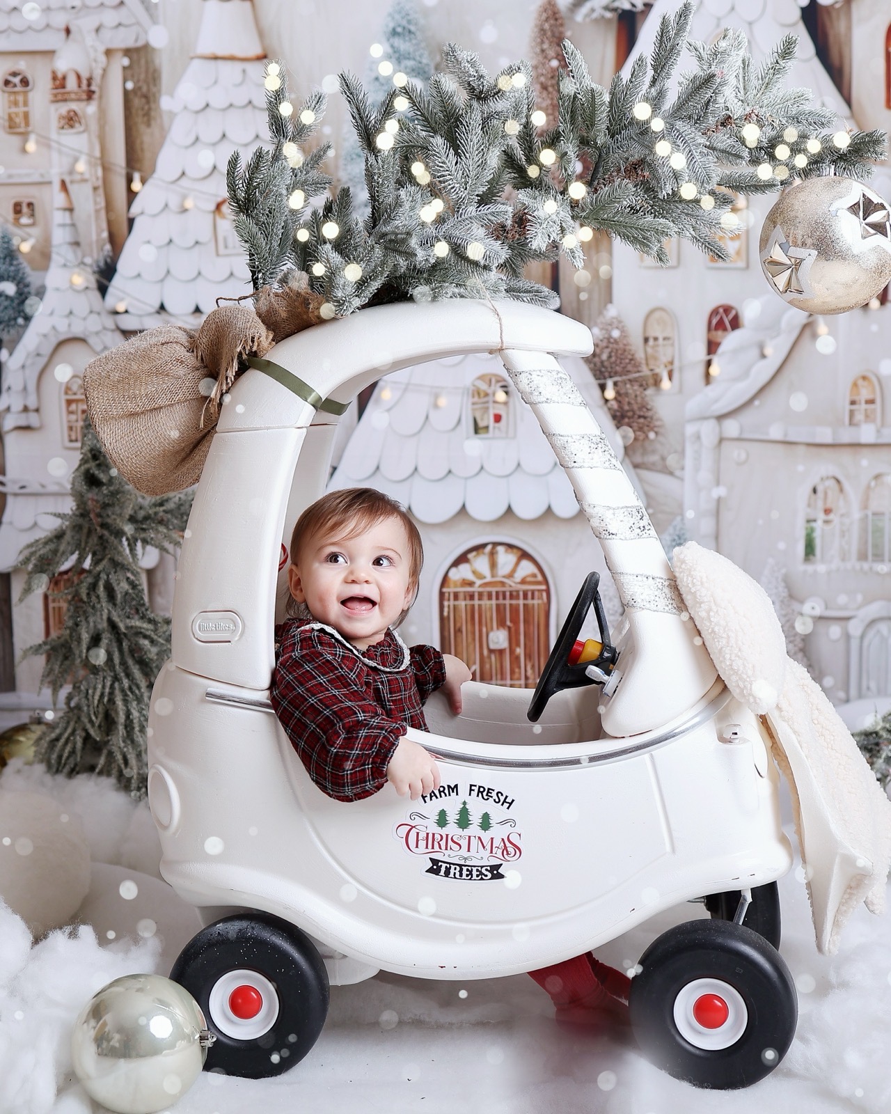 Toddler boy in a red plaid shirt sitting happily in a toy car under snowy pine branches