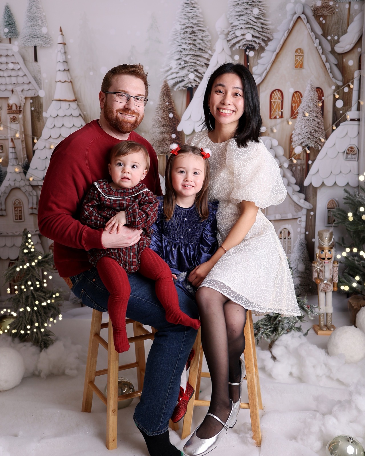Family of four sitting together on wooden stools, gingerbread village backdrop