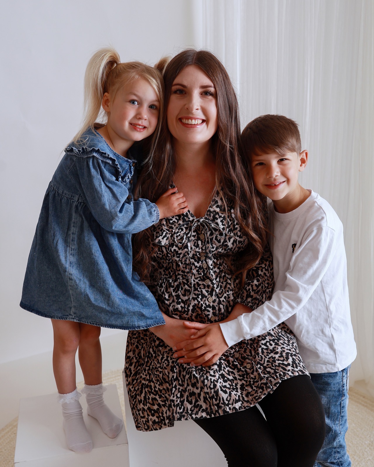 Mum in a leopard-print dress hugged closely by her daughter in a denim dress and son in a white top, both resting their hands on her shoulders