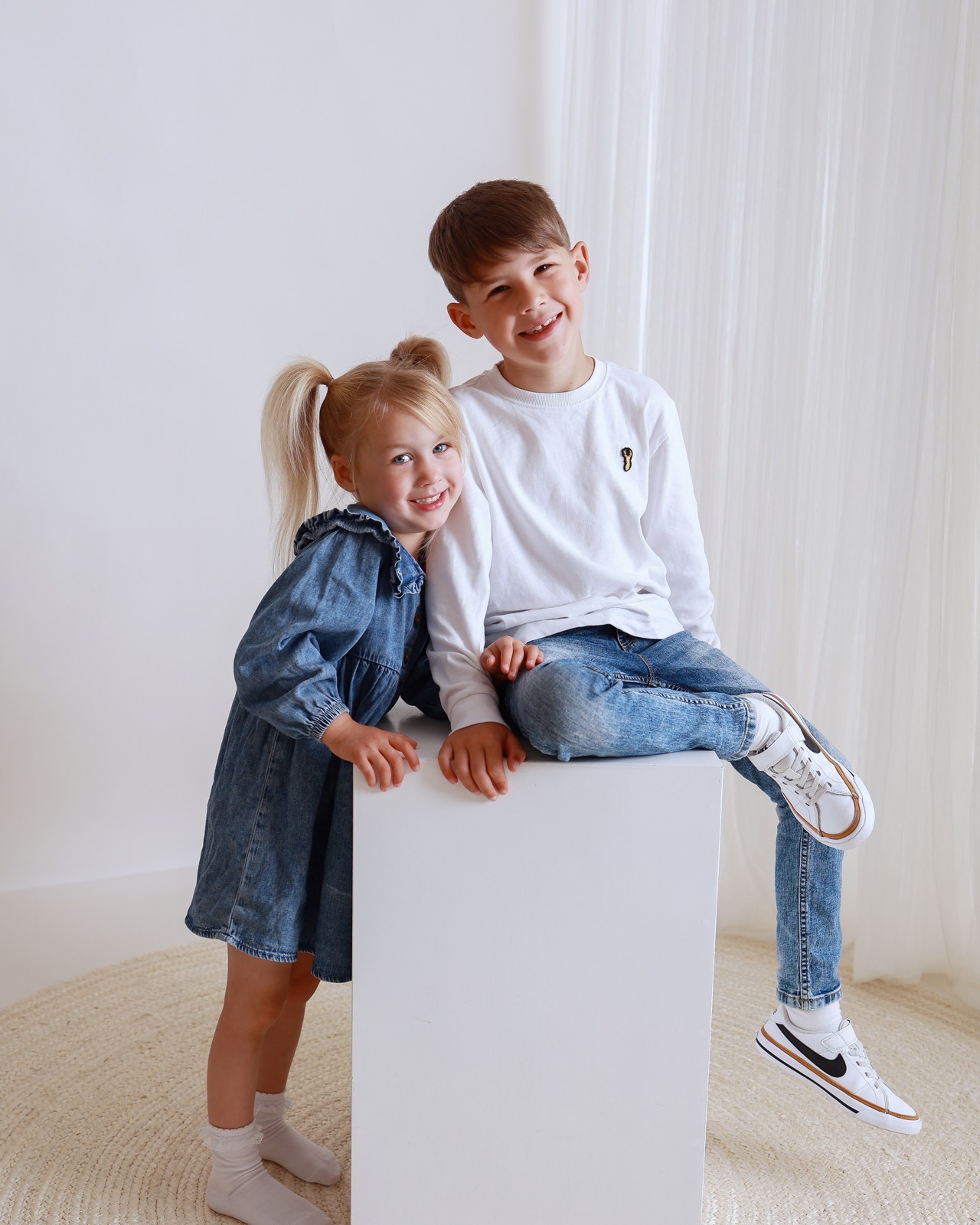 Brother and sister sitting together on a white cube, boy in a white top and jeans, girl in a denim dress