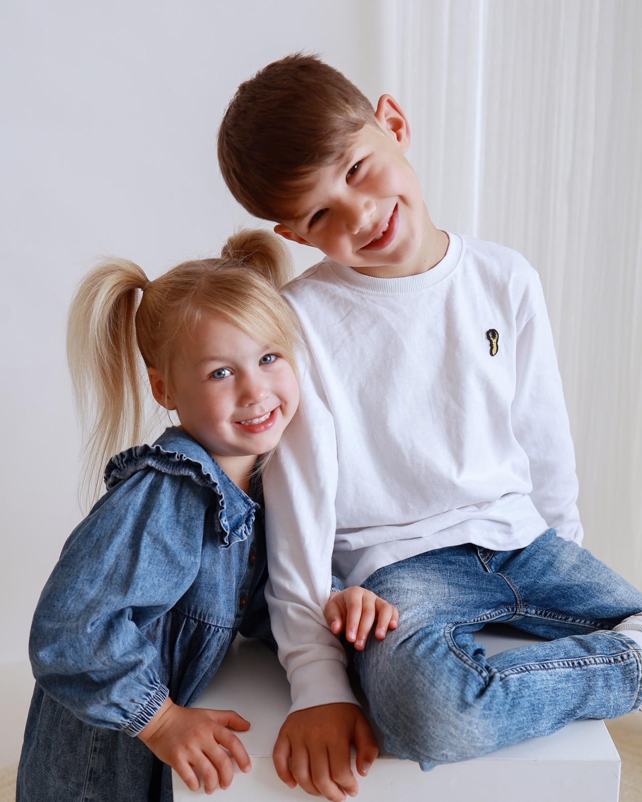 Brother and sister sitting close together, girl in a denim dress with her hair in pigtail buns, boy in a white top