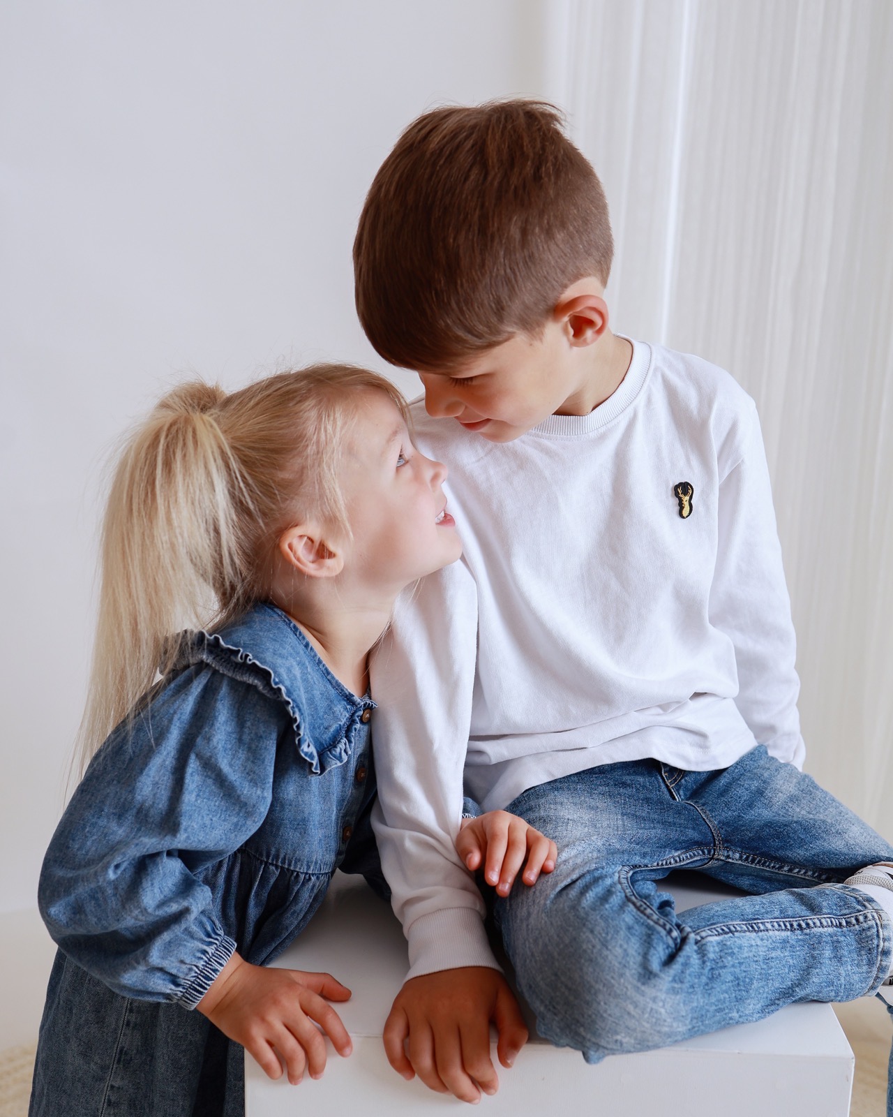 Brother and sister facing each other and smiling, girl in a denim dress, boy in a white top