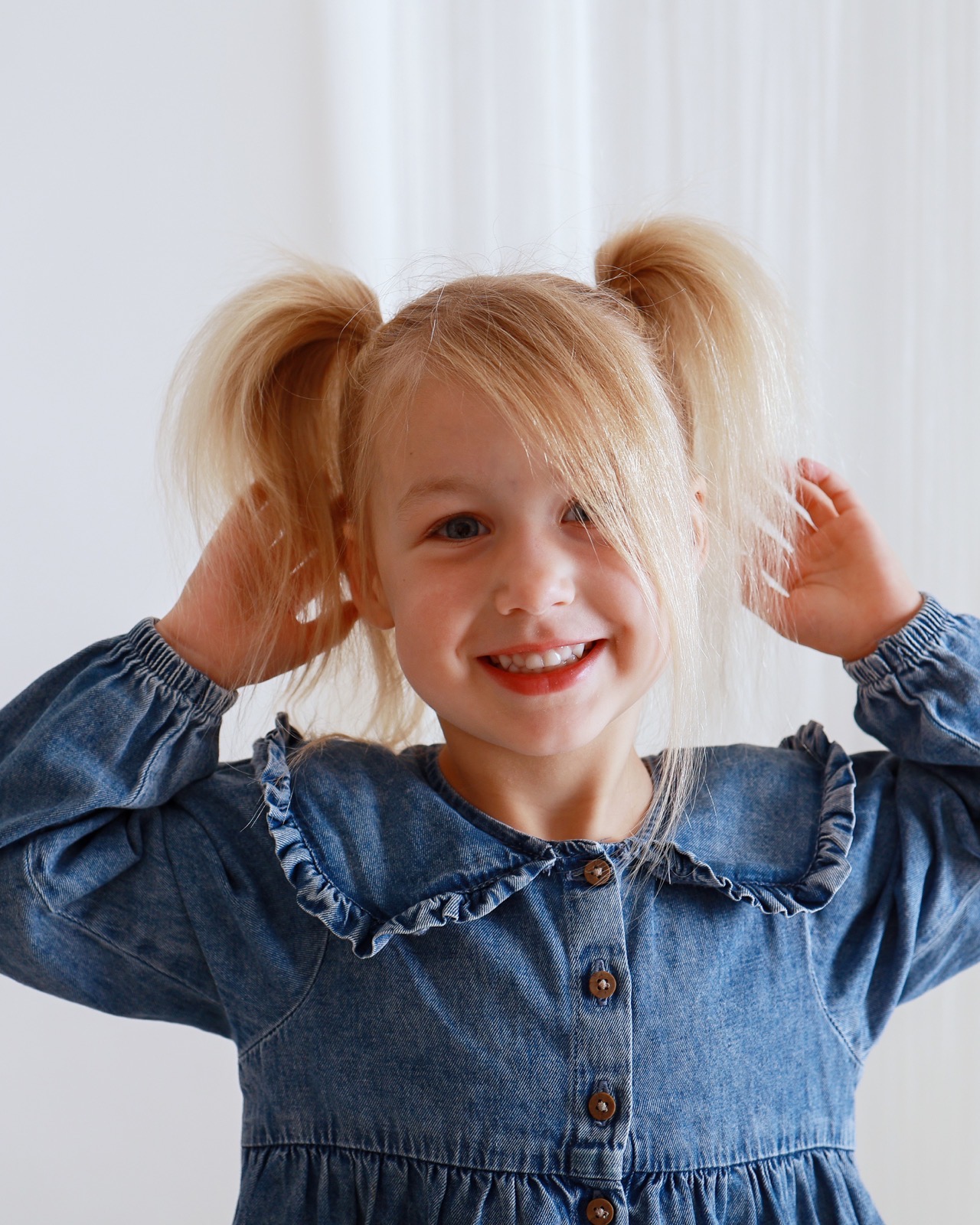 Close-up of a young girl playfully holding up her blonde pigtail buns, denim dress with a ruffled collar