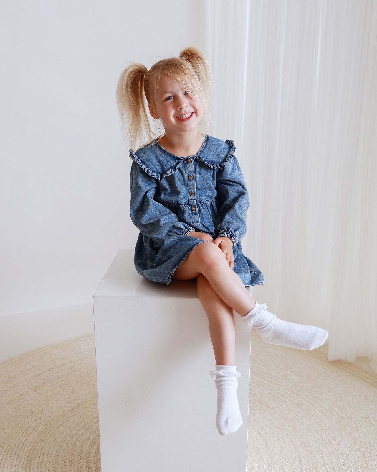 Girl sitting on a white cube with legs crossed, denim dress and white lace-trimmed socks