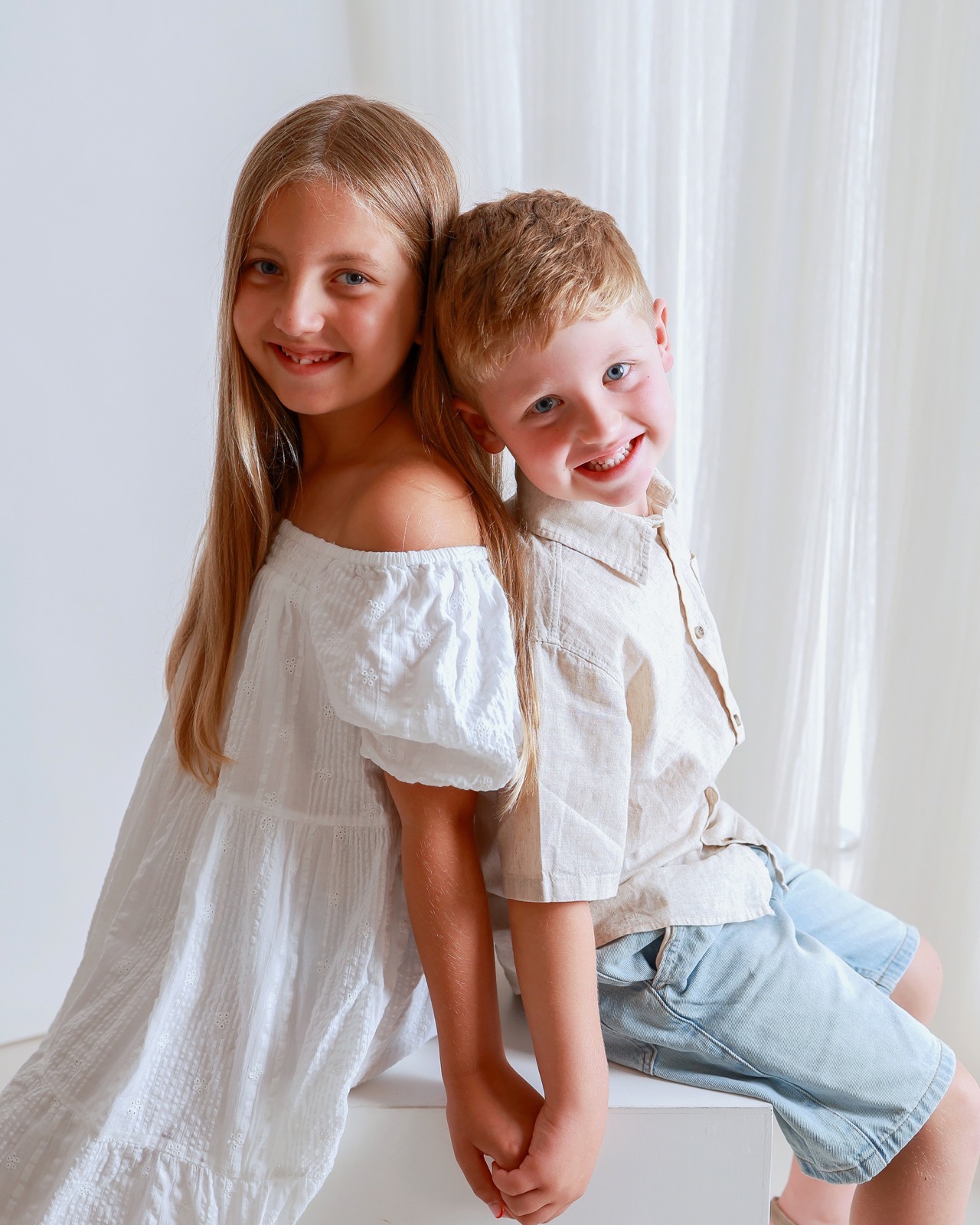 Older sister and younger brother sitting back to back, she in an off-the-shoulder white dress, he in a beige linen shirt