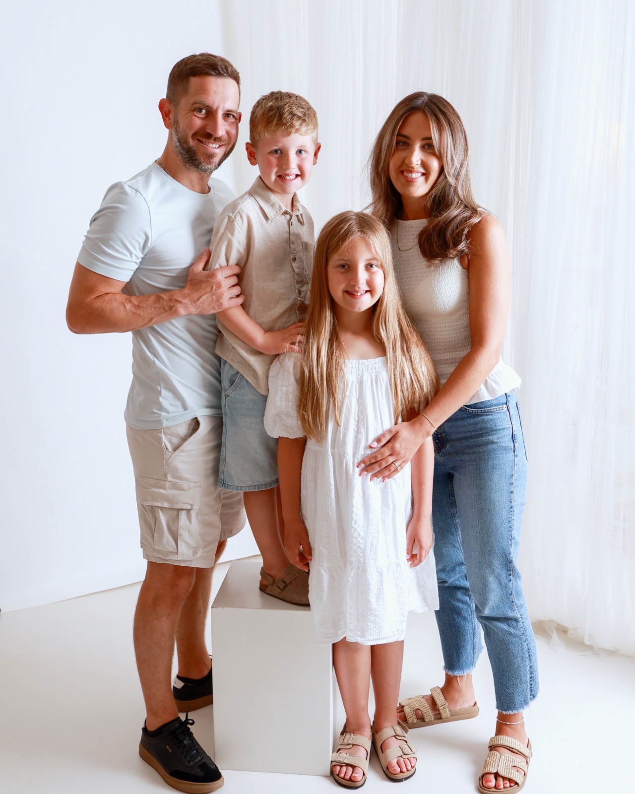 Family of four standing together, dad in a light blue top, boy in beige, girl in a white off-shoulder dress, mum in cream