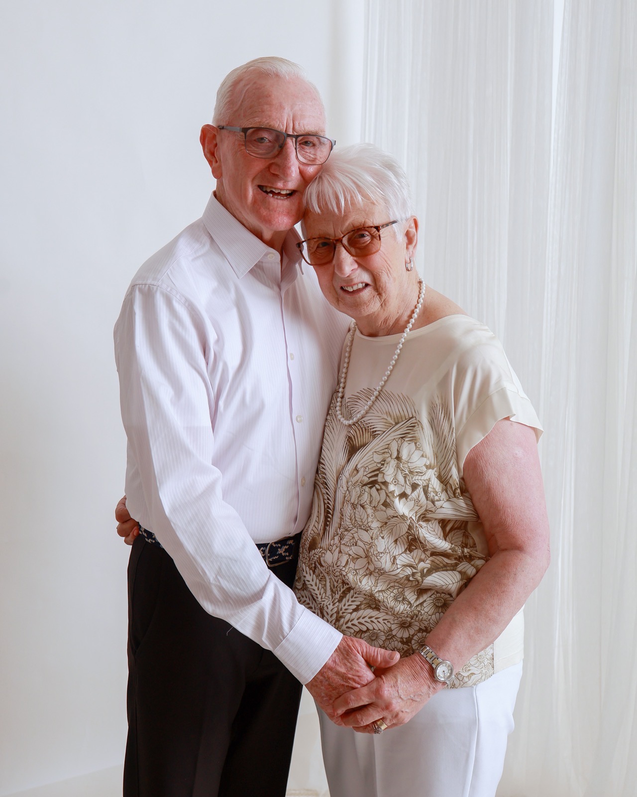 Elderly couple embracing, him in a pale pink shirt and her in a cream floral top with a pearl necklace