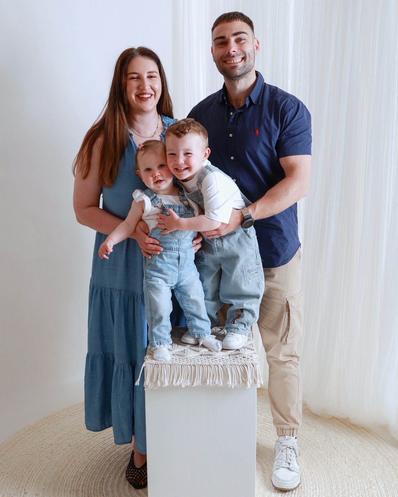 Young family of four, mum in a teal dress and dad in a navy shirt, with two toddlers in denim dungarees standing on a stool