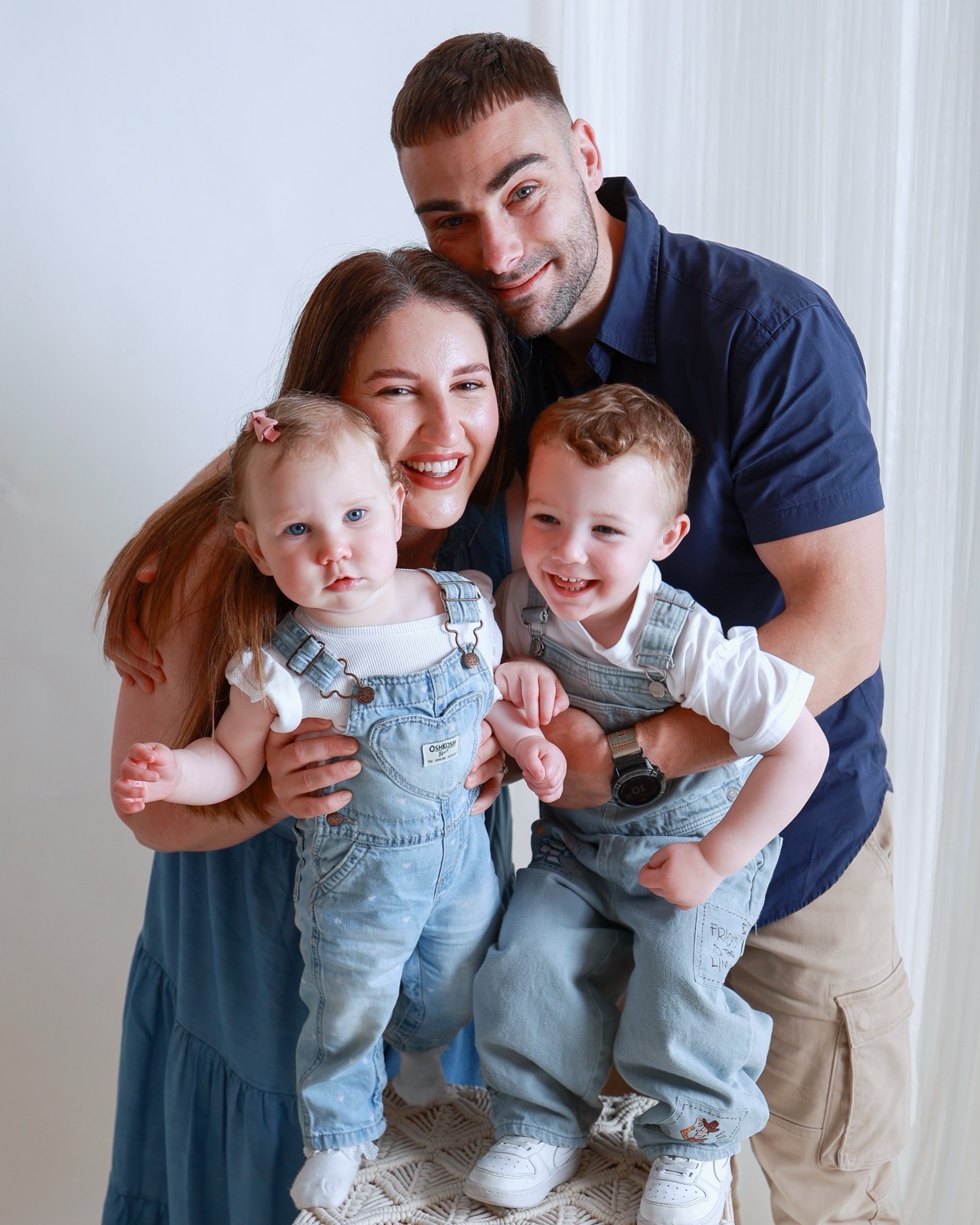 Young family embracing closely, baby girl and toddler boy in denim dungarees held between their parents
