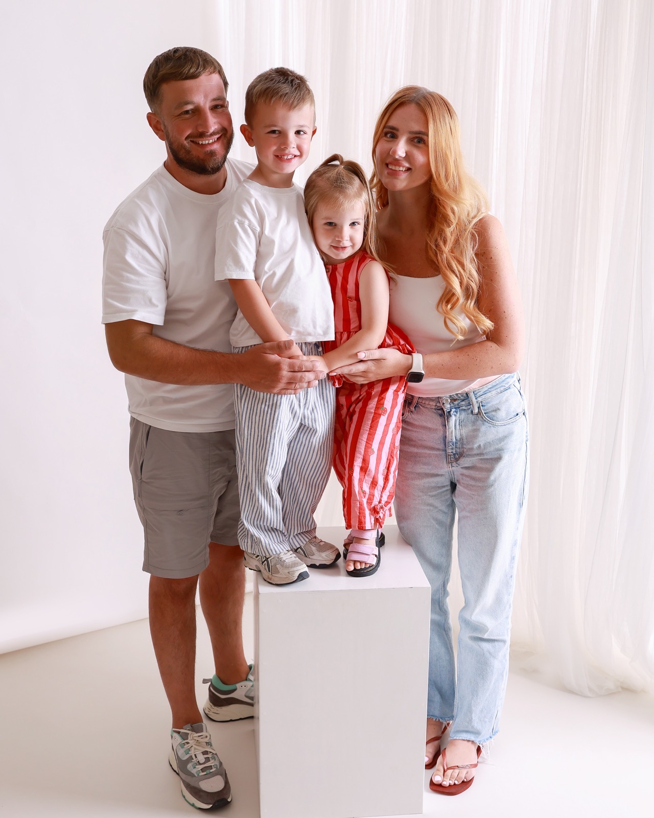Family of four holding hands together, dad in grey shorts, boy in striped trousers, girl in a red strawberry-print outfit, mum in denim