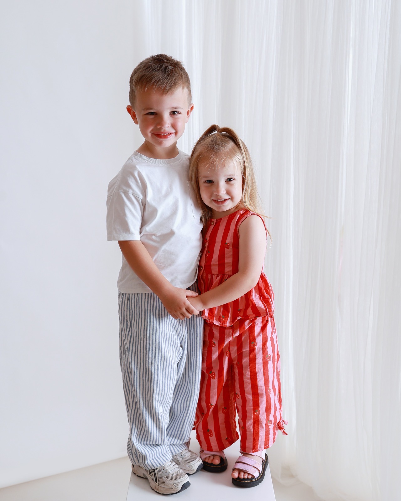 Brother and sister holding hands and hugging, boy in striped trousers, girl in a red strawberry-print outfit