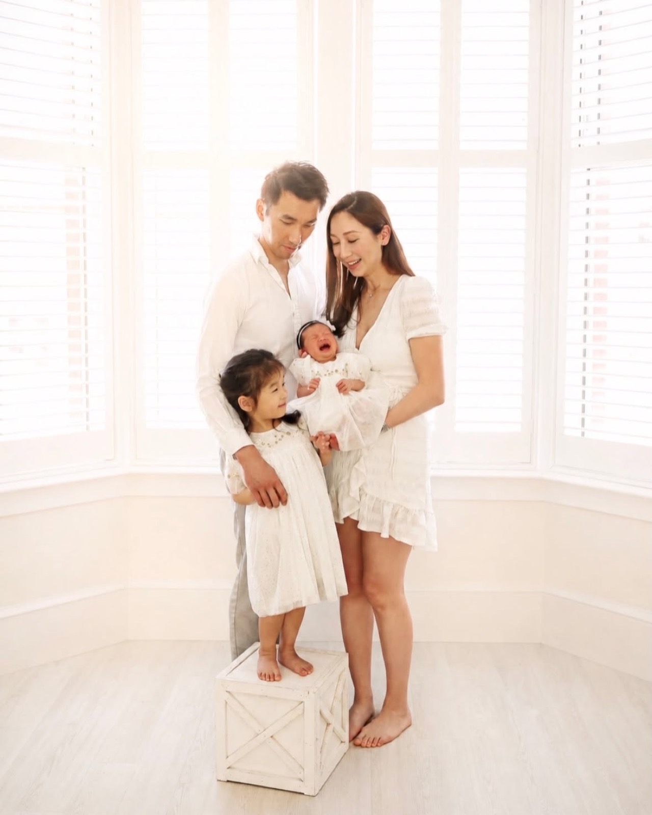 Family of four by a bright window, dad and mum smiling at their crying newborn while their young daughter looks on, all in white