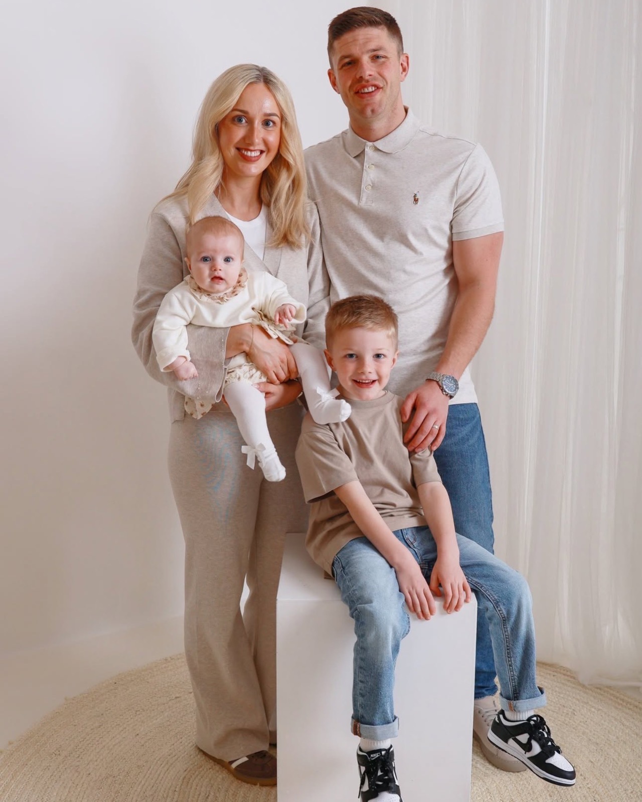 Family of four in matching oatmeal tones, mum holding baby girl, dad and older son sitting together