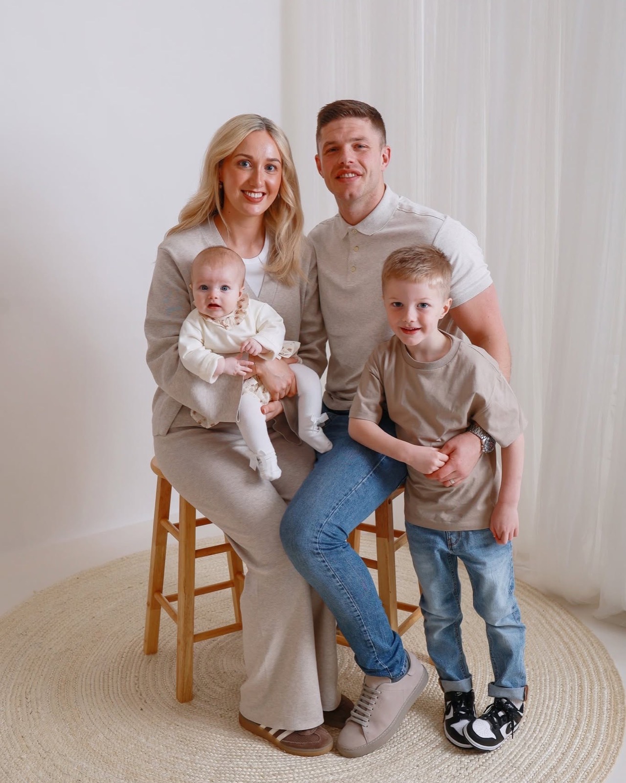 Family in matching oatmeal tones standing together, boy sitting on a wooden stool, dad holding the baby