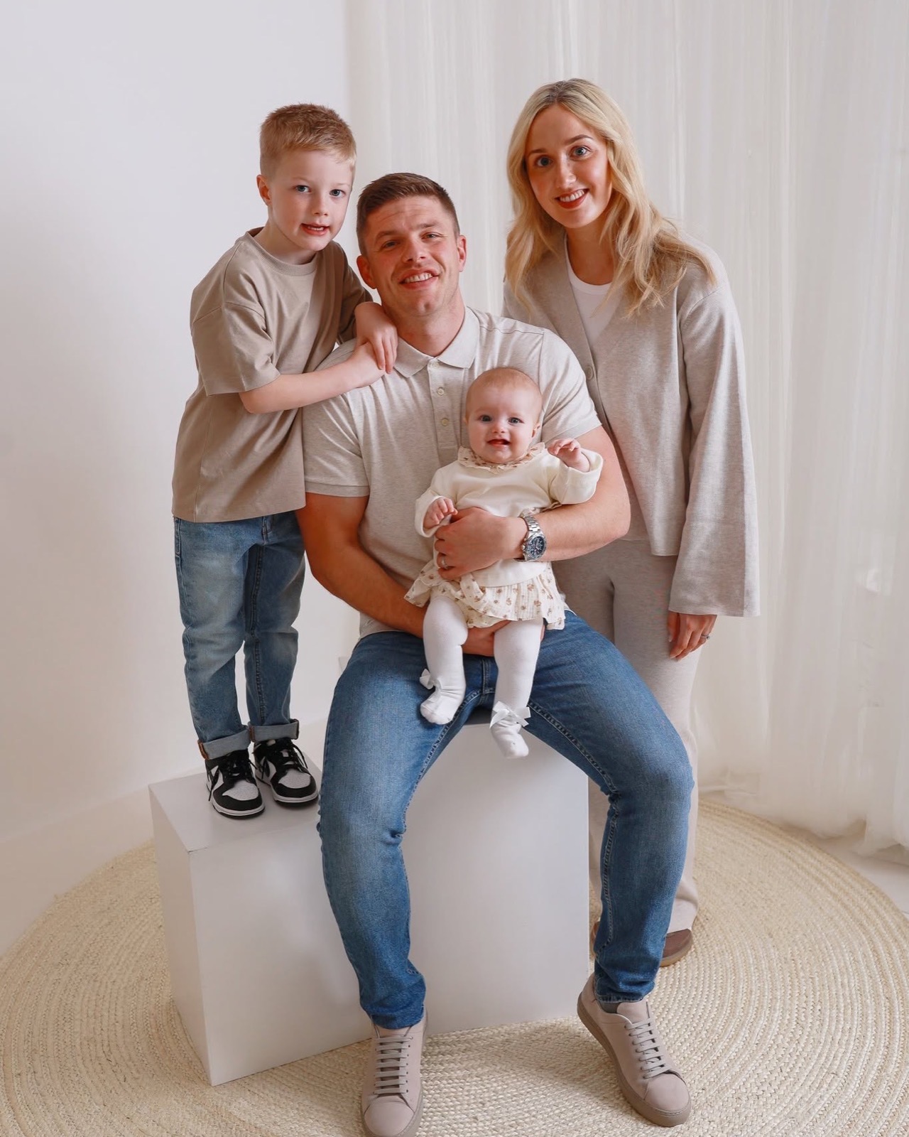 Family in matching oatmeal tones gathered closer, boy standing on a plinth beside his parents and baby sister