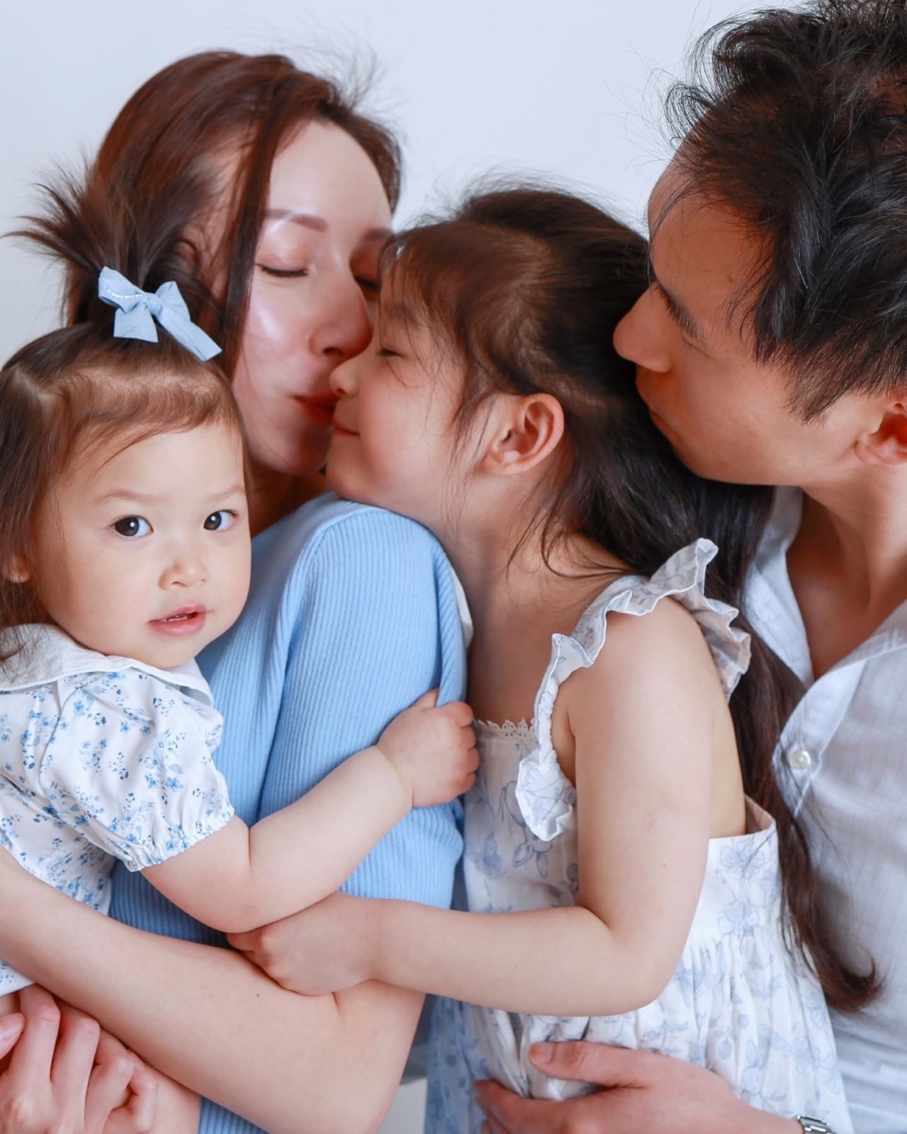 Mum and dad each kissing one of their two young daughters, one in a blue cardigan and one in a floral dress