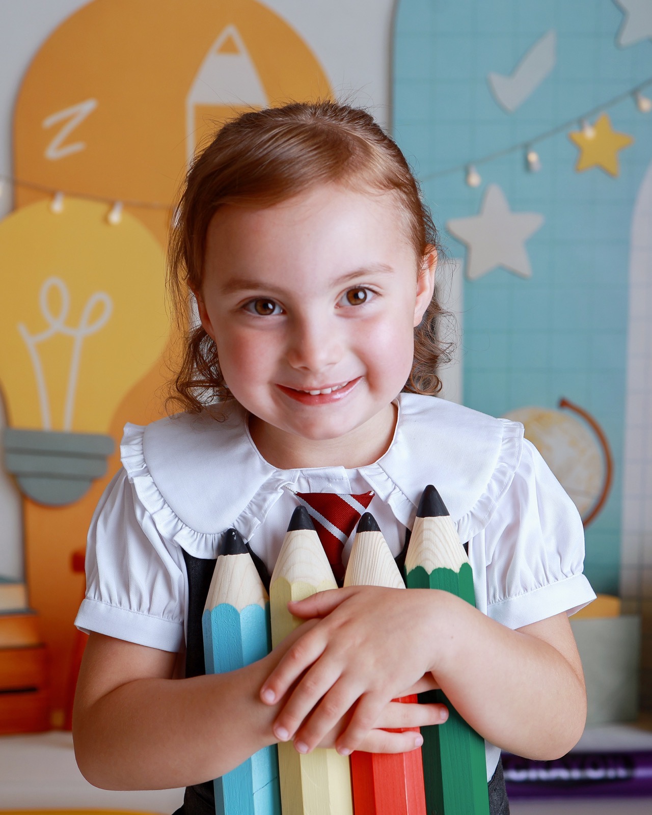 Girl smiling and hugging a bundle of oversized colored pencils, wearing a white blouse with a striped tie, classroom-themed backdrop