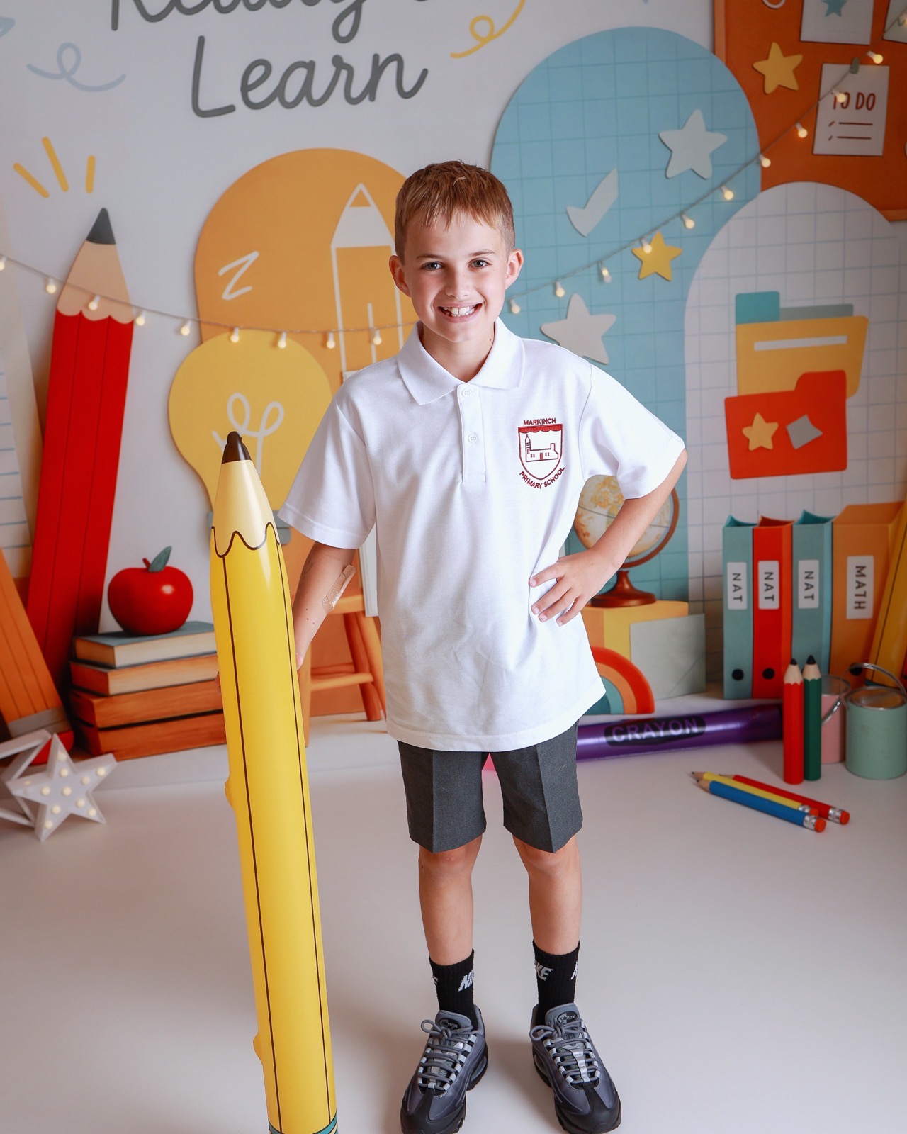 Boy standing beside a giant inflatable pencil prop, hand on hip, in a white polo with an embroidered school crest and navy shorts