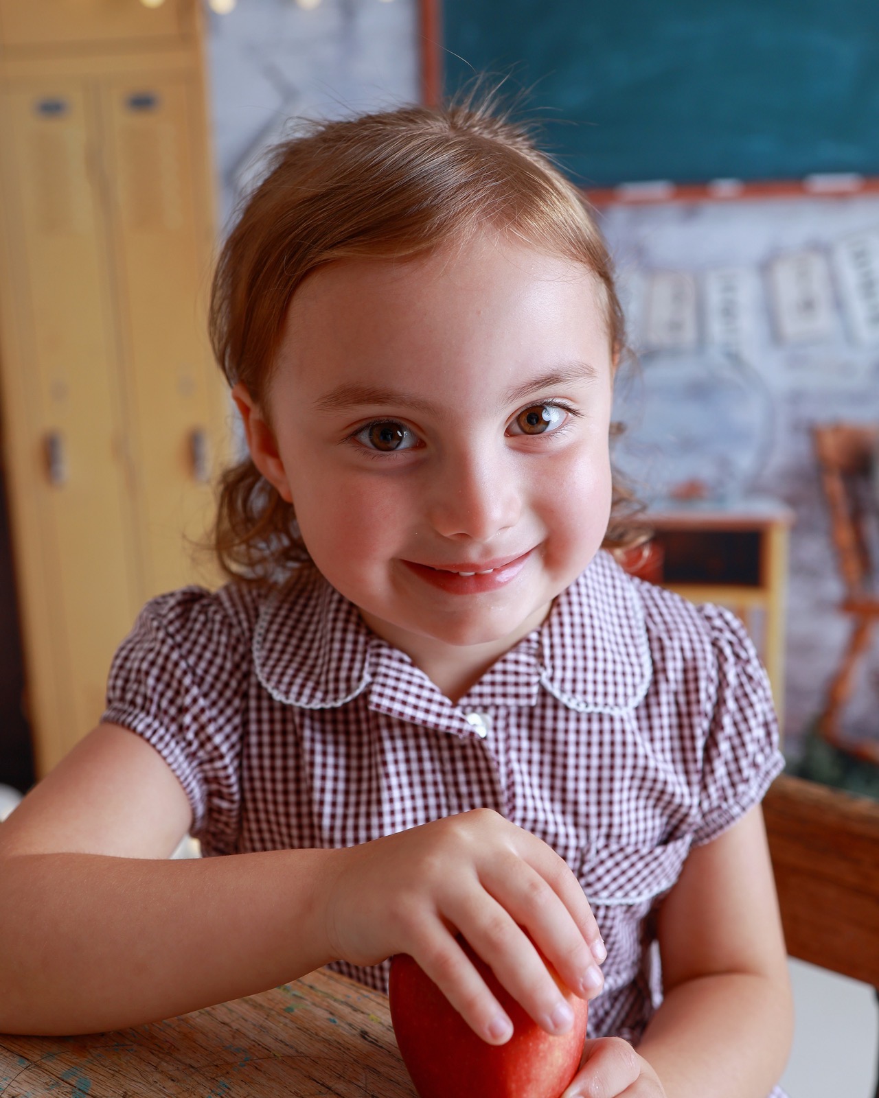 Close-up of a girl in a red gingham dress holding a red apple at a desk, chalkboard in the background