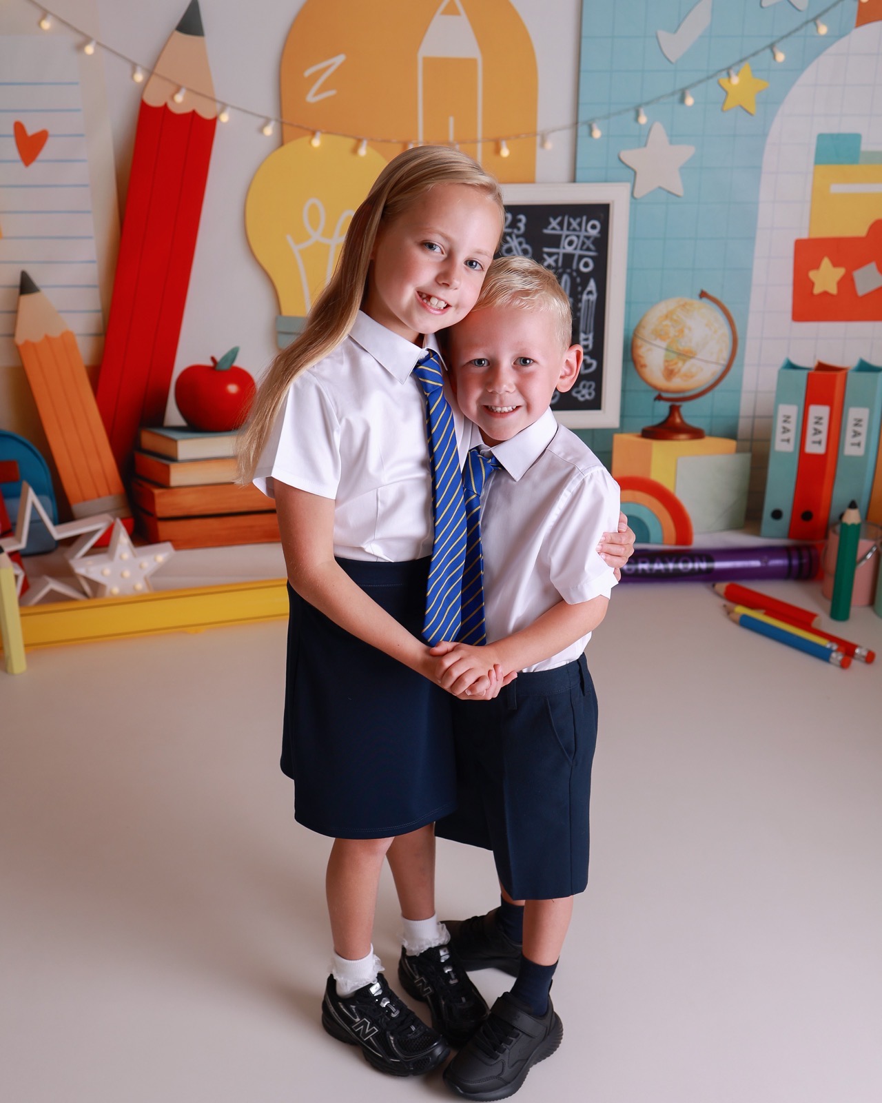 Older sister hugging her younger brother, both in white shirts and striped ties, classroom-themed backdrop