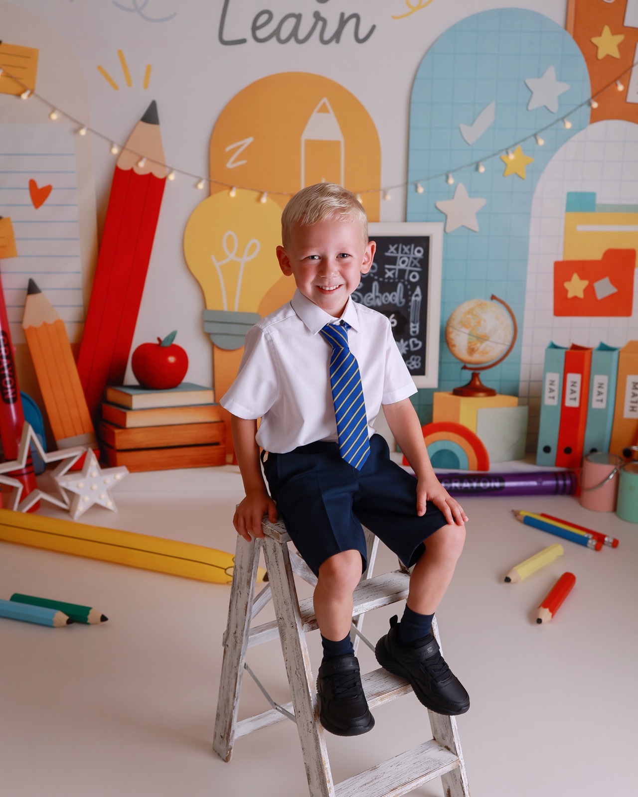Boy sitting on a small step stool, white shirt and blue striped tie, navy shorts, smiling