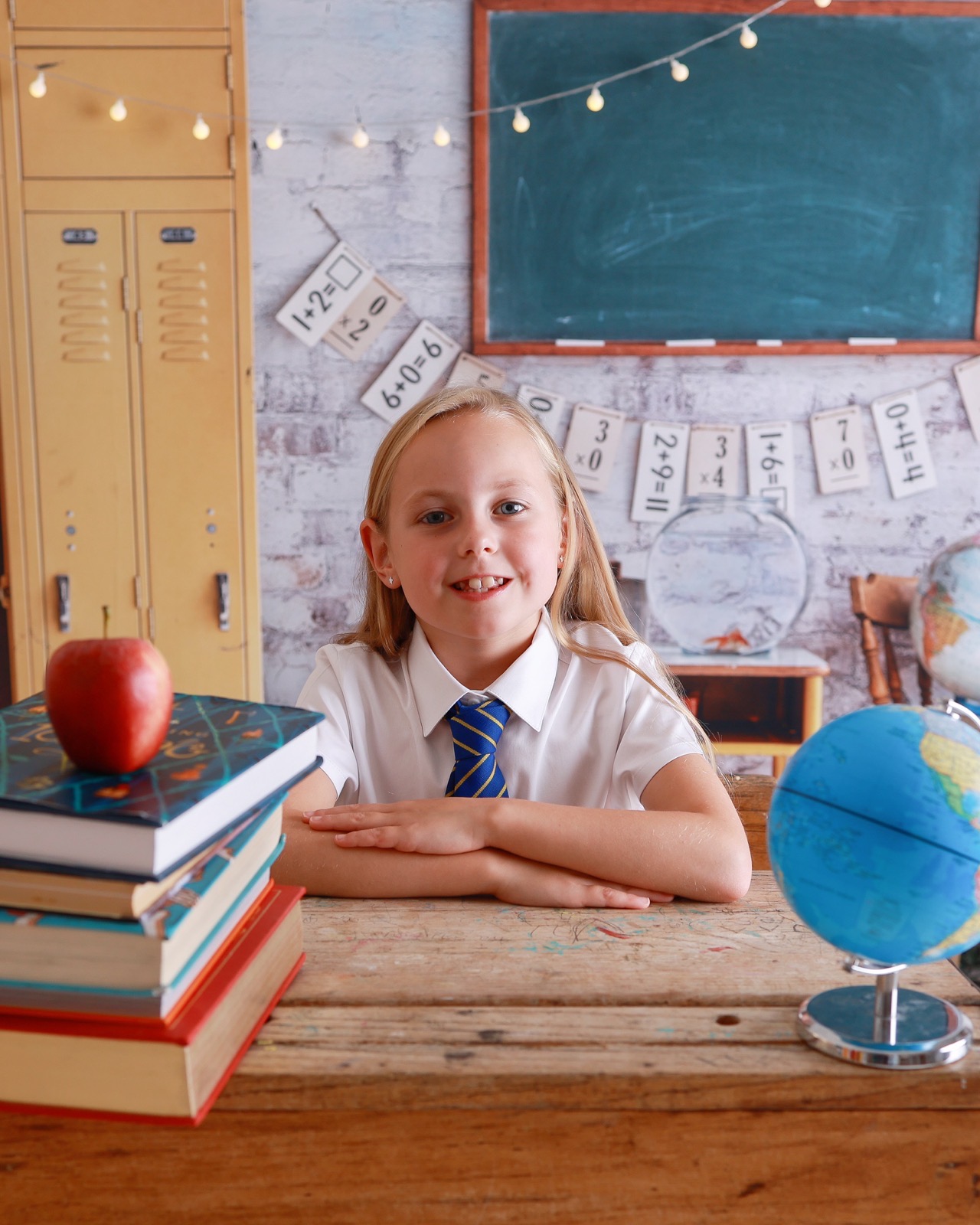 Girl sitting at a vintage school desk with stacked books, an apple and a globe, striped tie and glasses