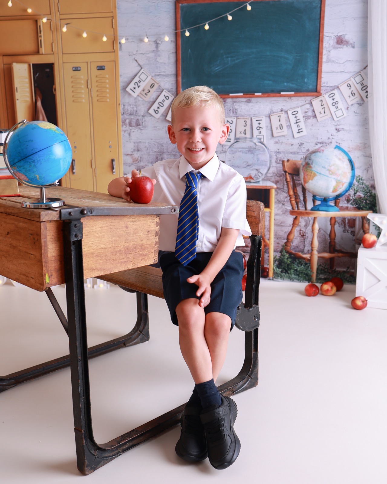 Boy sitting at a vintage school desk holding a red apple, globe beside him, chalkboard behind