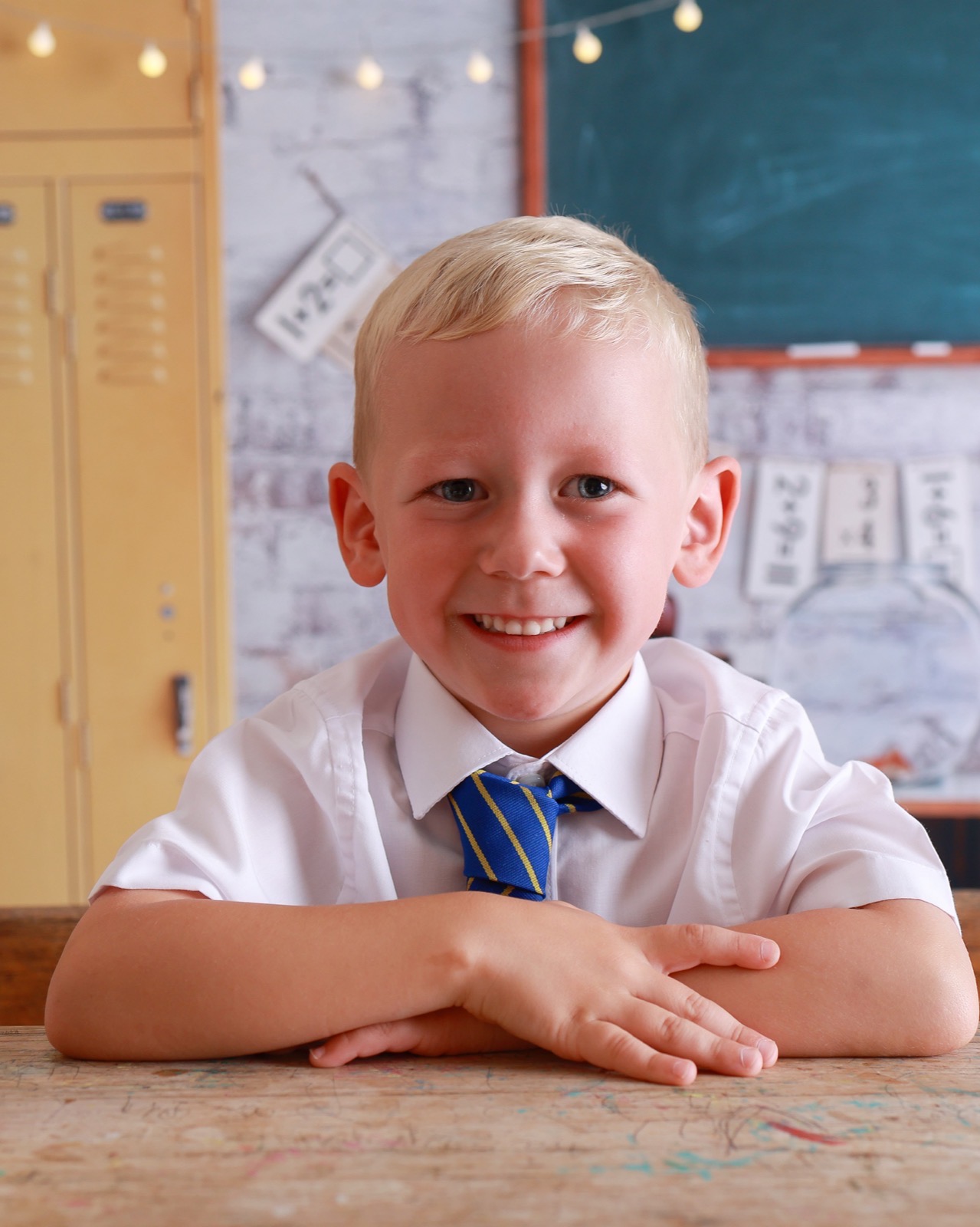 Close-up of a smiling boy leaning on a desk with arms crossed, striped tie, chalkboard backdrop
