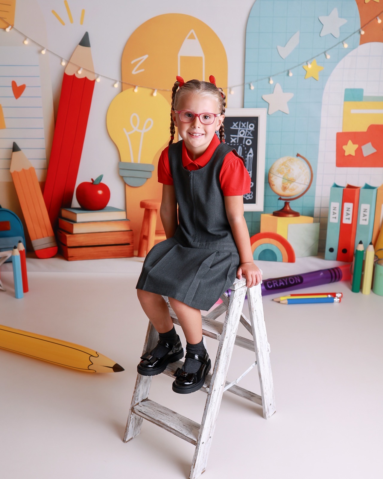 Girl standing on a step stool in a grey pinafore and red top, pigtails and glasses