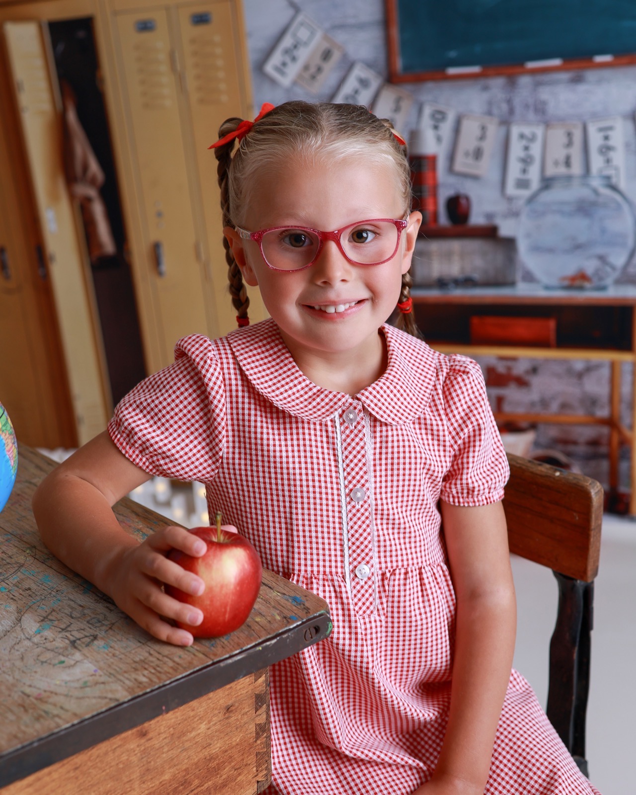 Close-up of a smiling girl in a red gingham dress and glasses, holding a red apple