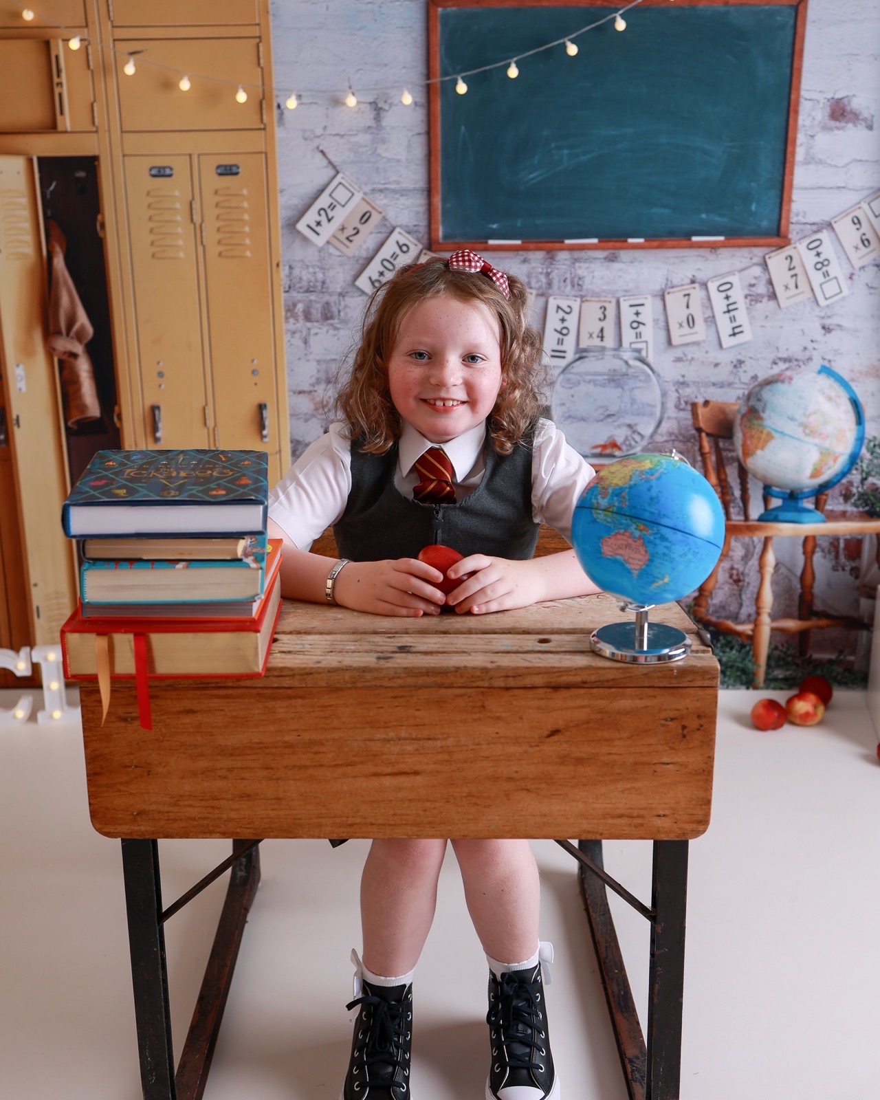 Girl sitting at a desk with books, an apple and a globe, grey pinafore and striped tie, curly hair
