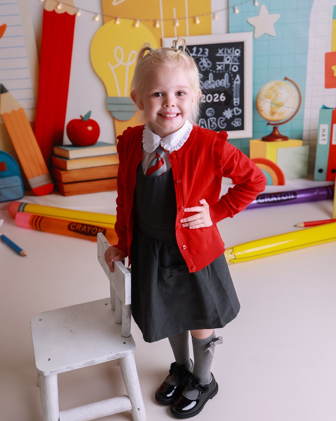 Girl standing beside a small white step stool, red cardigan and grey pinafore, hand on hip
