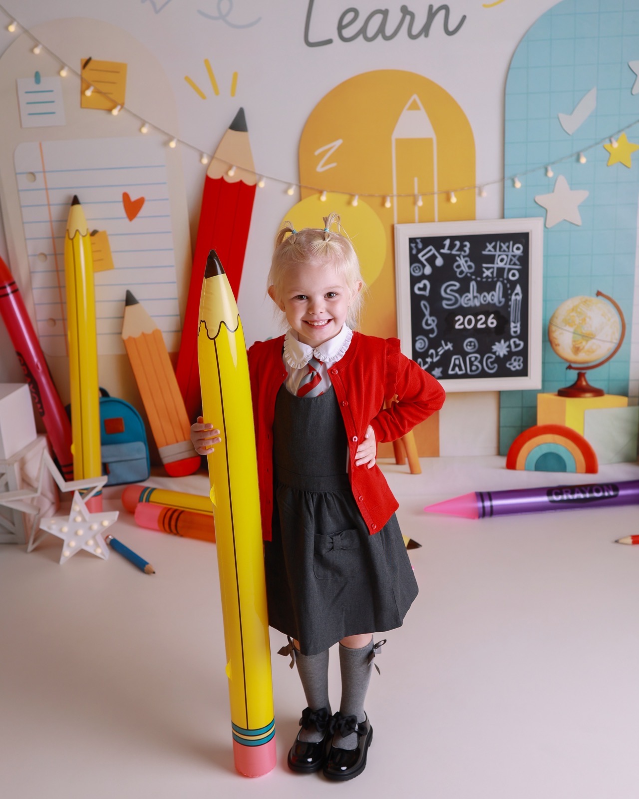 Girl standing beside a giant inflatable pencil prop, red cardigan, grey pinafore and striped tie