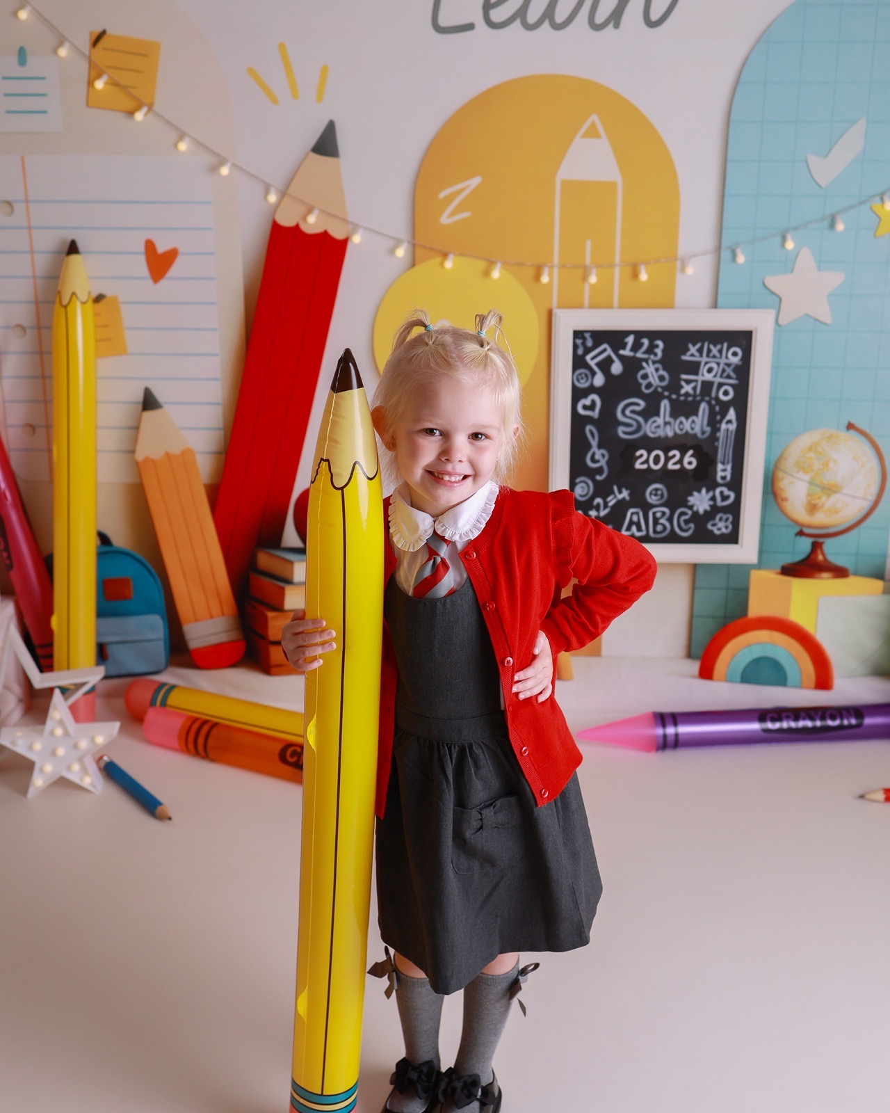 Girl leaning against a giant inflatable pencil prop, red cardigan and pinafore, smiling
