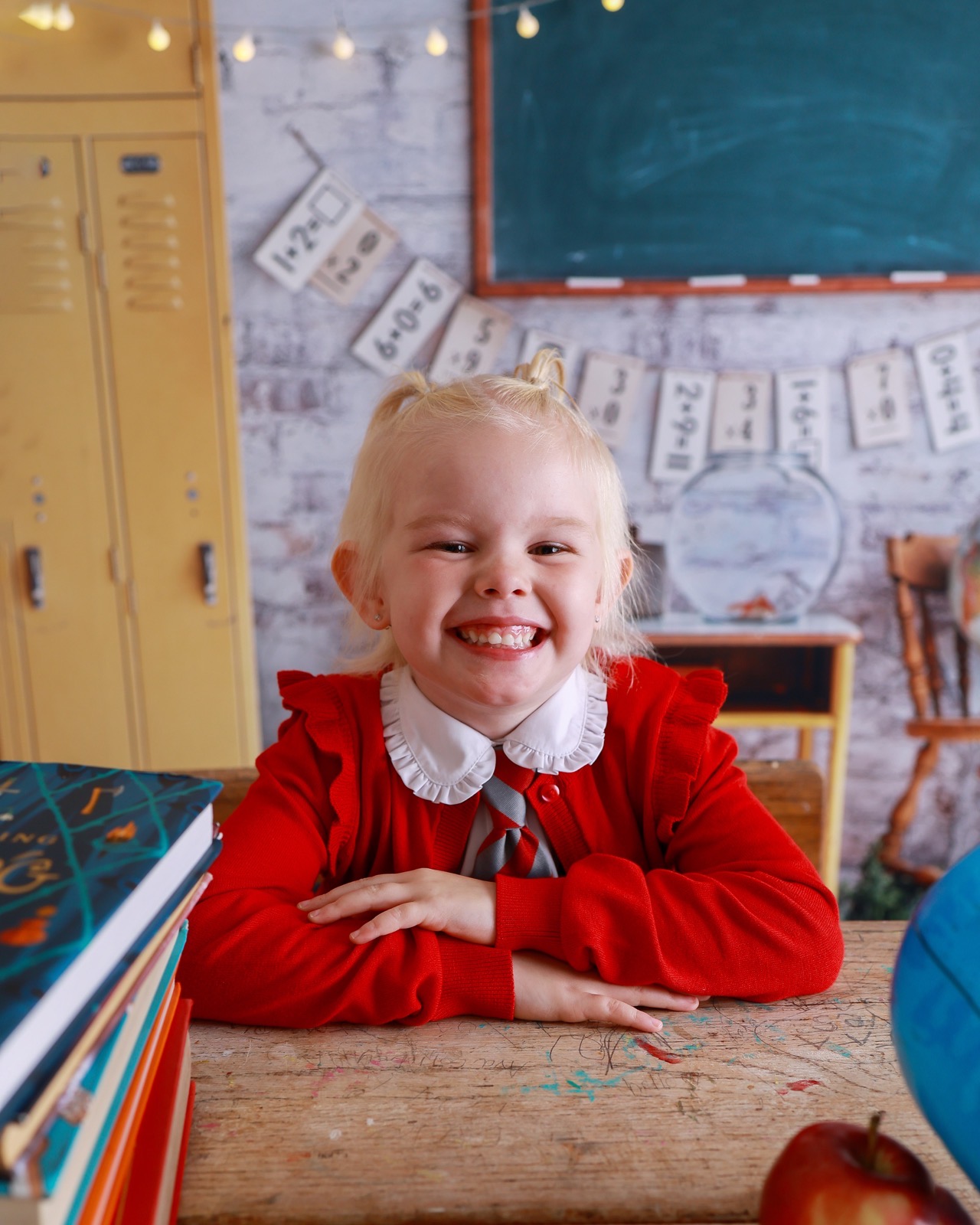 Close-up of a smiling girl resting her chin on her hands at a desk, red cardigan, hair in two buns