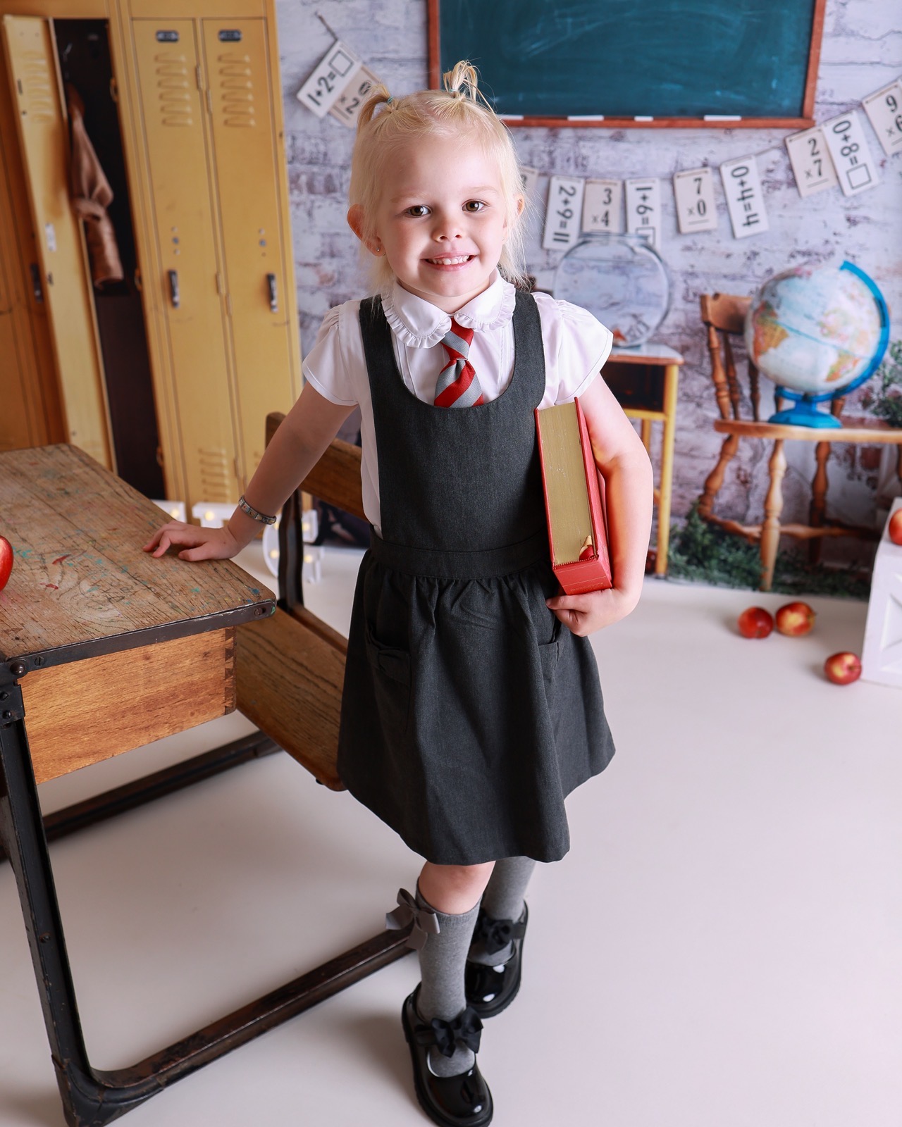 Girl sitting on a wooden desk with legs dangling, grey pinafore, striped tie and black shoes with bows