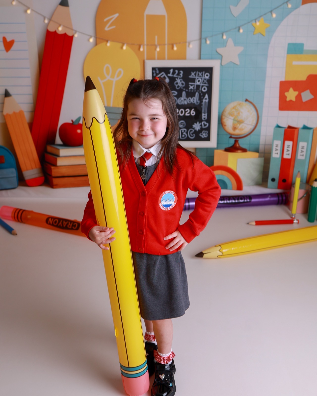 Girl holding a giant inflatable pencil prop, red cardigan with an embroidered crest, grey skirt