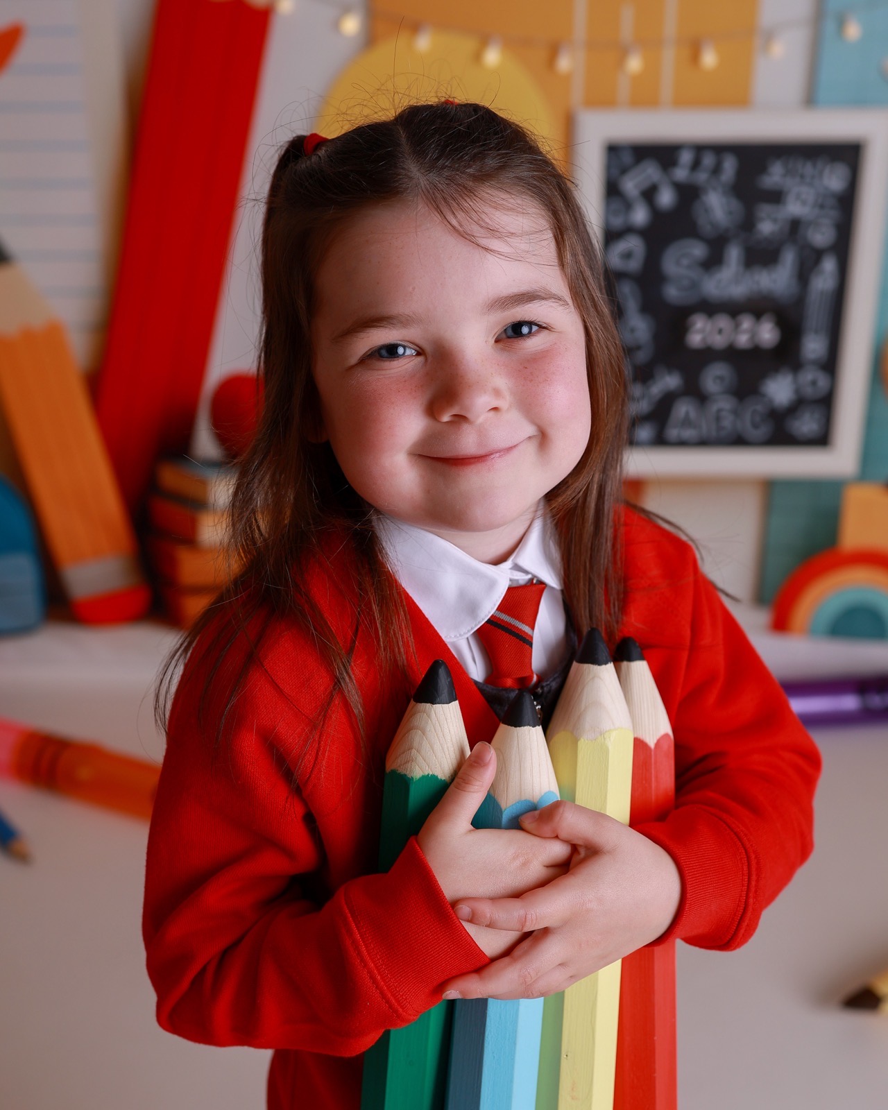 Close-up of a girl in a red cardigan holding a bundle of giant colored pencils, striped tie