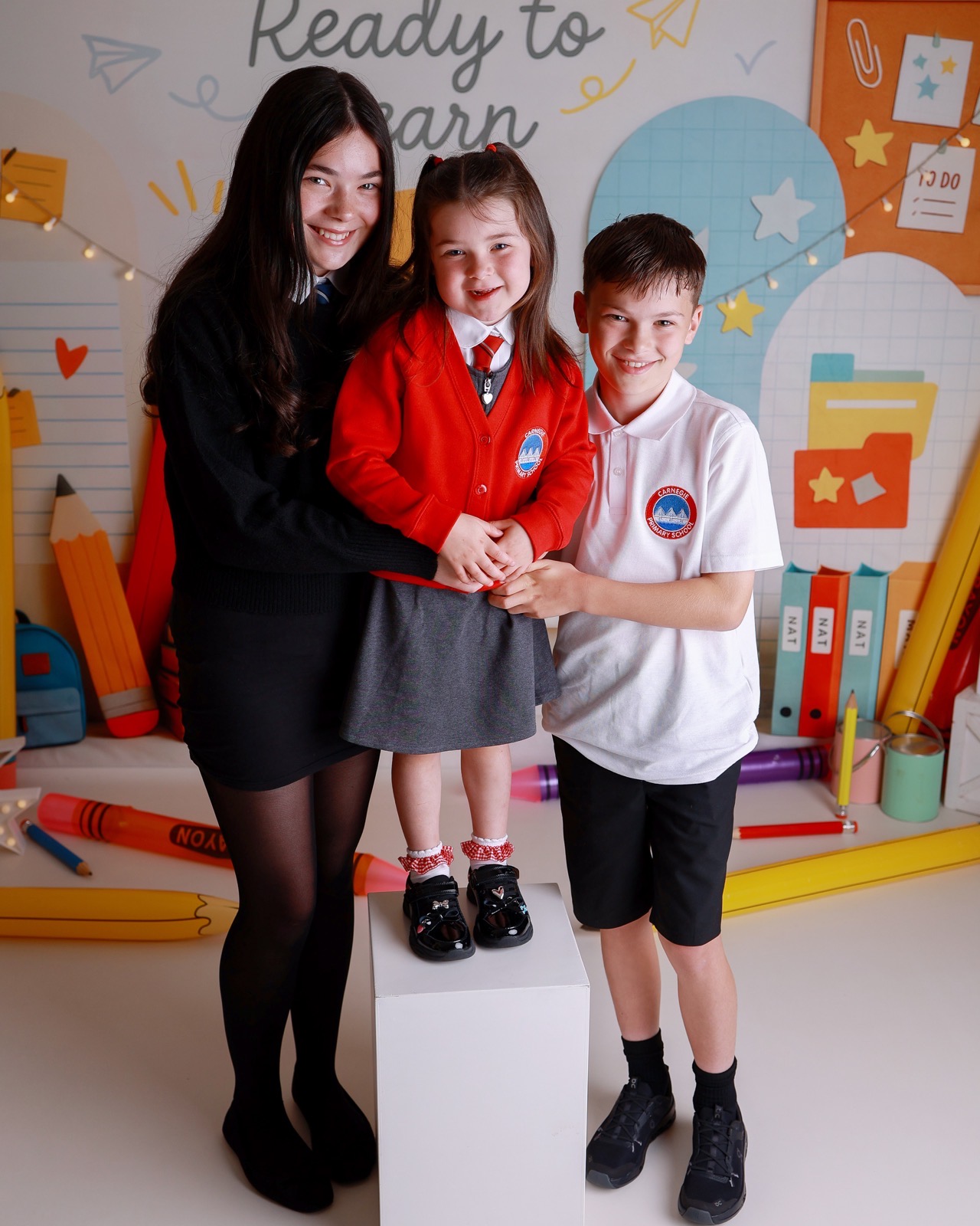 Three siblings standing together in school uniforms, the youngest perched on a small white plinth