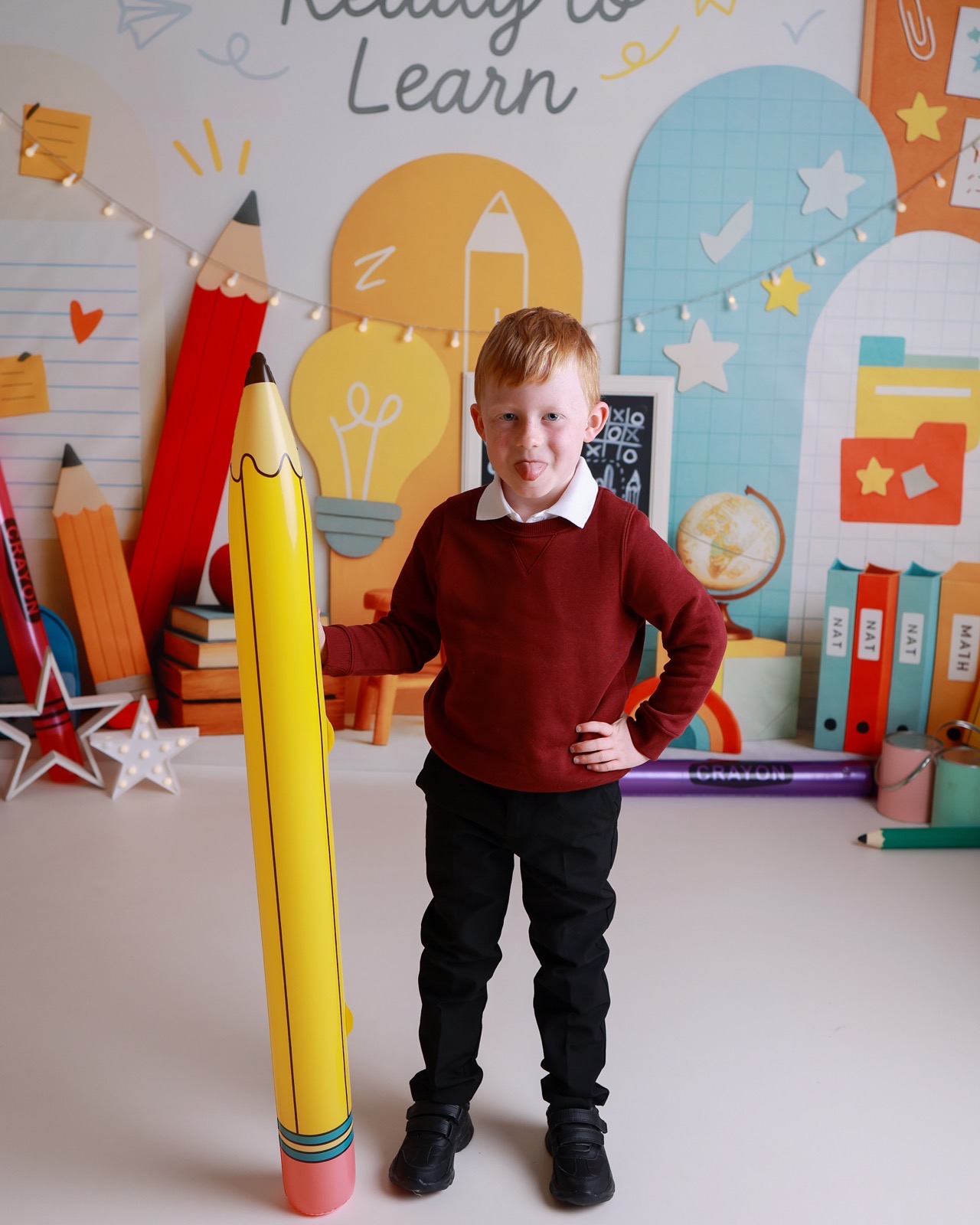 Boy pulling a silly face with his tongue out beside a giant inflatable pencil prop, maroon jumper