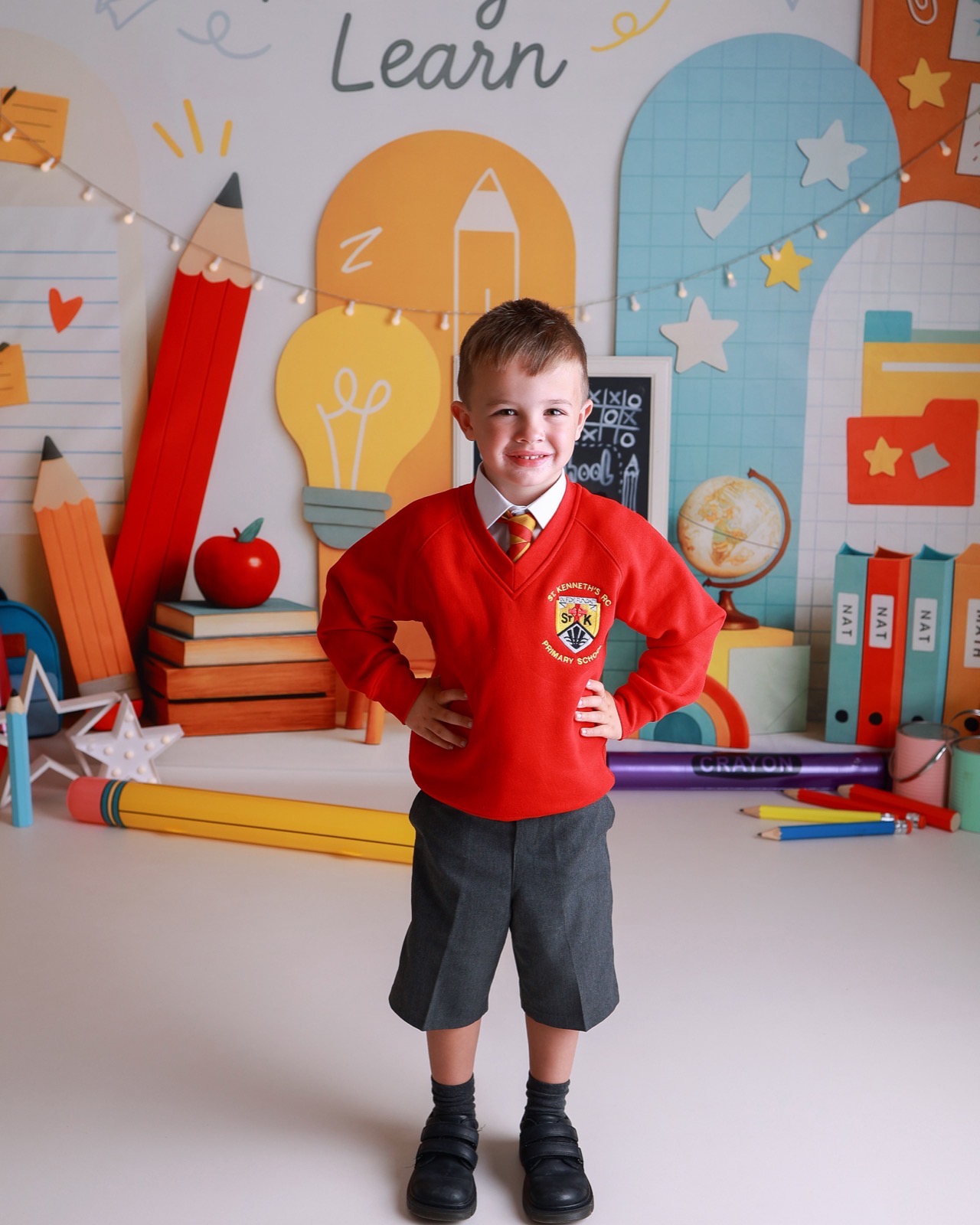 Boy standing with hands on hips in a red V-neck jumper with an embroidered crest and grey shorts