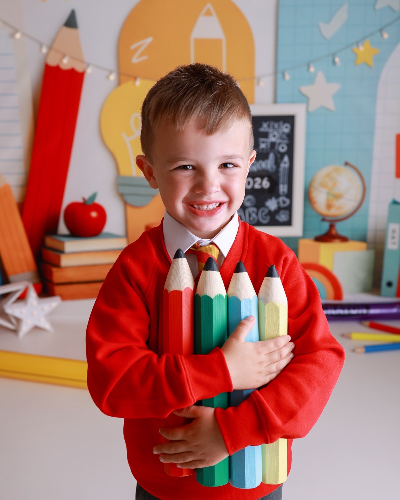 Boy hugging a bundle of giant colored pencils, red jumper and striped tie, gap-toothed smile