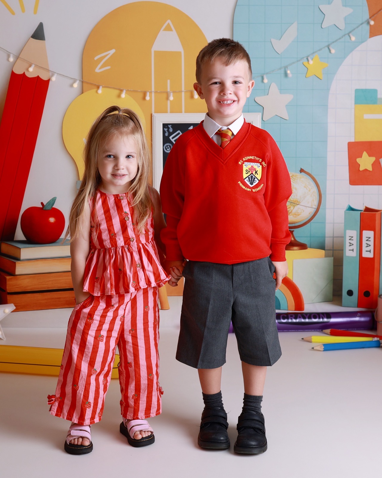 Younger sister in a striped strawberry-print outfit holding hands with her brother in a red school jumper